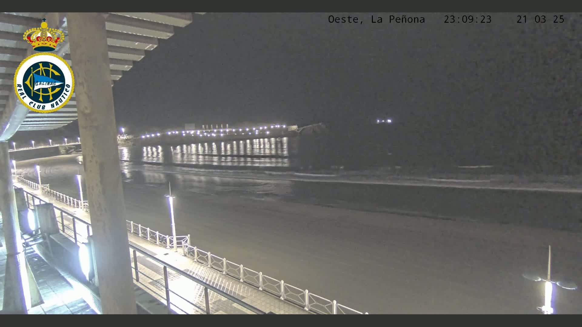 A nighttime view of a pier extending into a dark ocean with gentle waves lapping at the shore, under a clear sky.