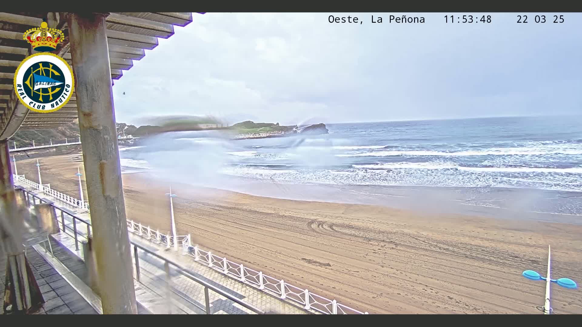 A sandy beach with moderate waves under an overcast sky is seen from a high vantage point overlooking a boardwalk.
