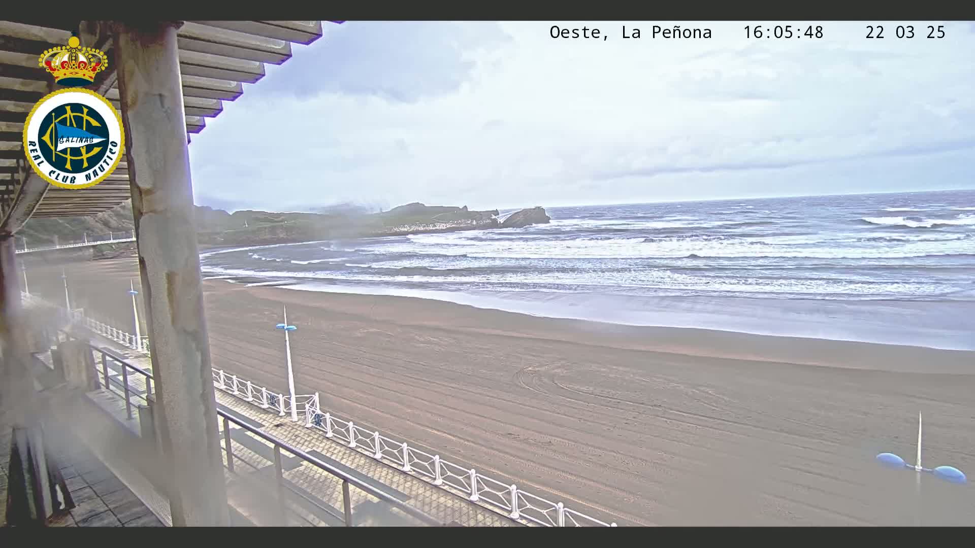 A sandy beach with moderate waves under a cloudy sky is viewed from a covered balcony.