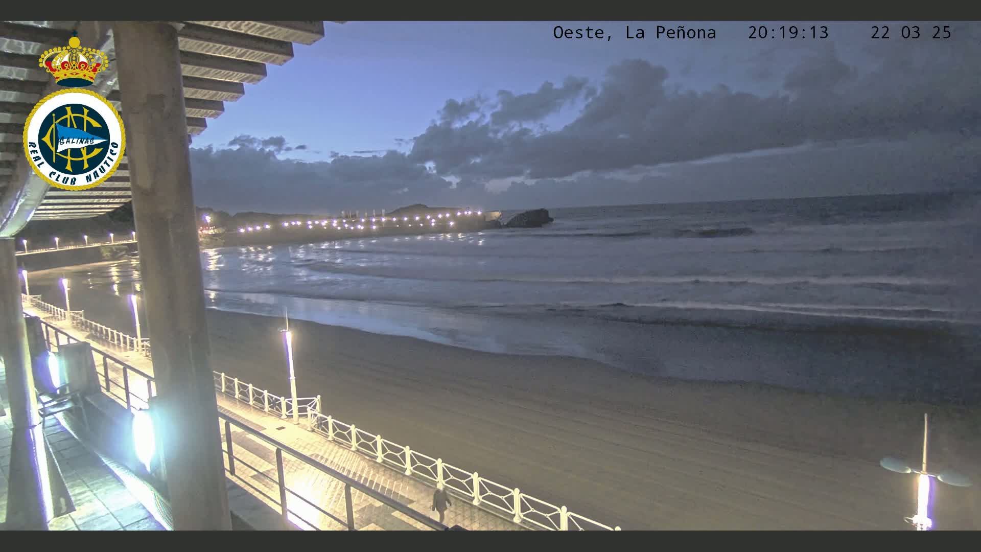 A sandy beach at twilight, under a partly cloudy sky, is seen from an elevated position, showing a boardwalk with lights extending along the shore and gentle waves rolling onto the beach.