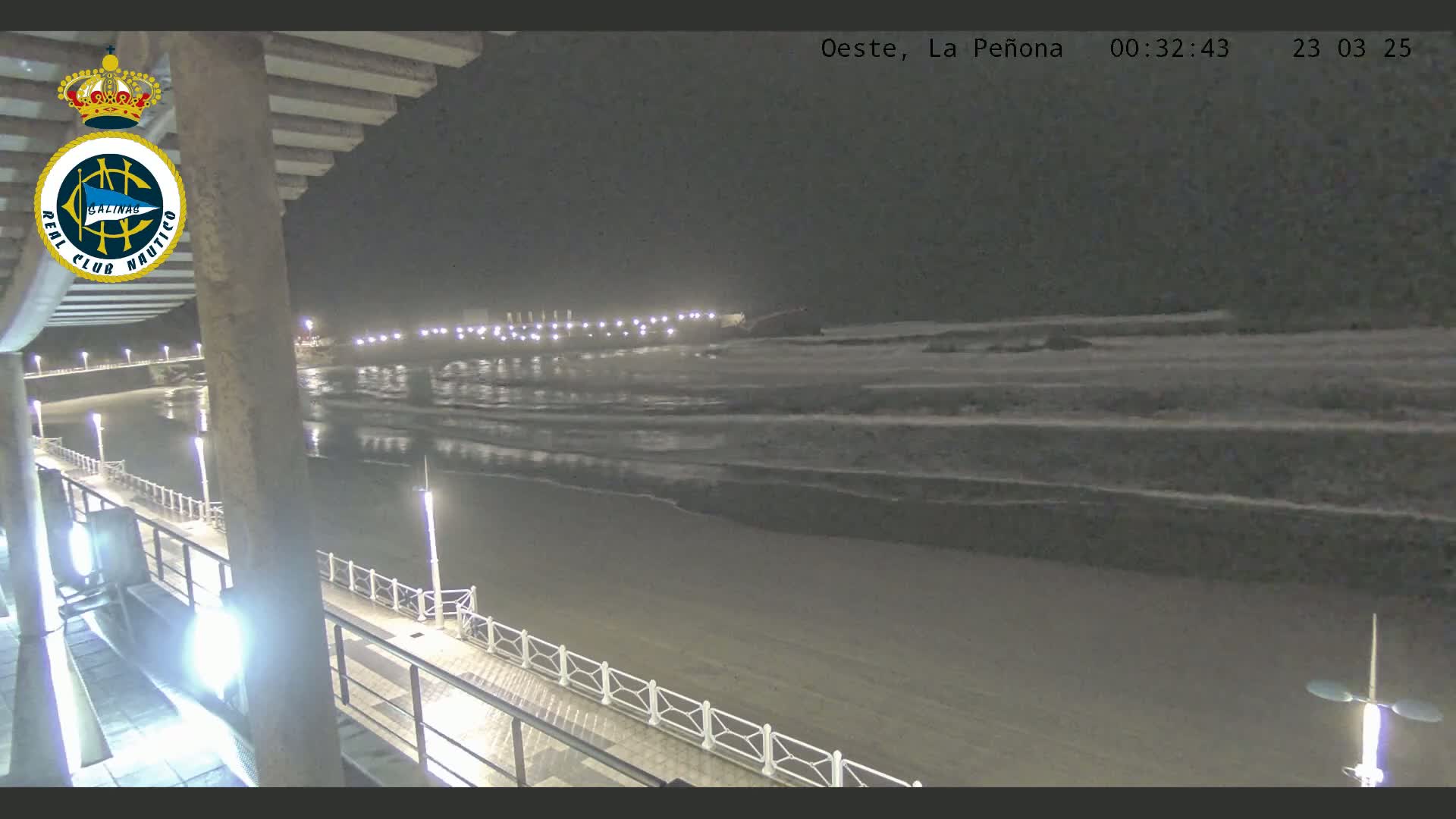 A nighttime view of a beach with gentle waves lapping the shore, illuminated by lights along a pier and boardwalk.