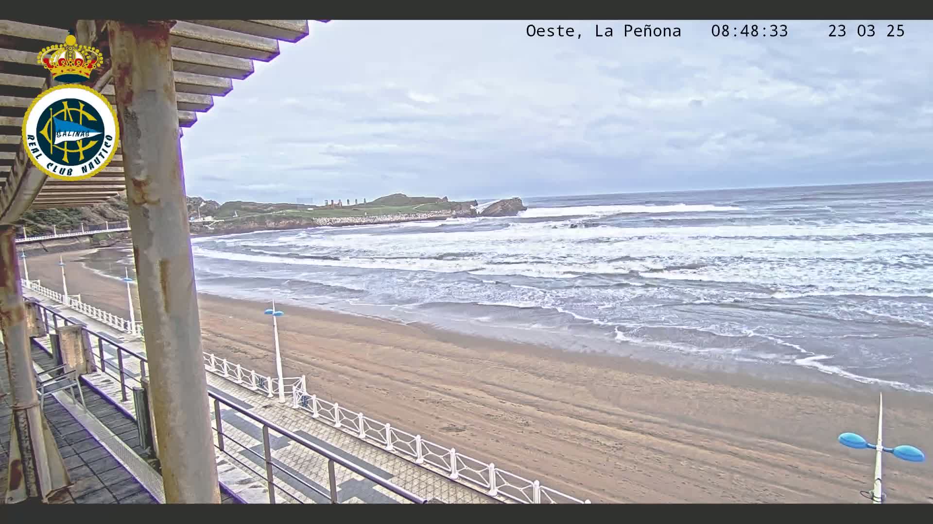 A sandy beach with moderate waves under a cloudy sky is viewed from an elevated position, overlooking a paved walkway and a structure with a roof.