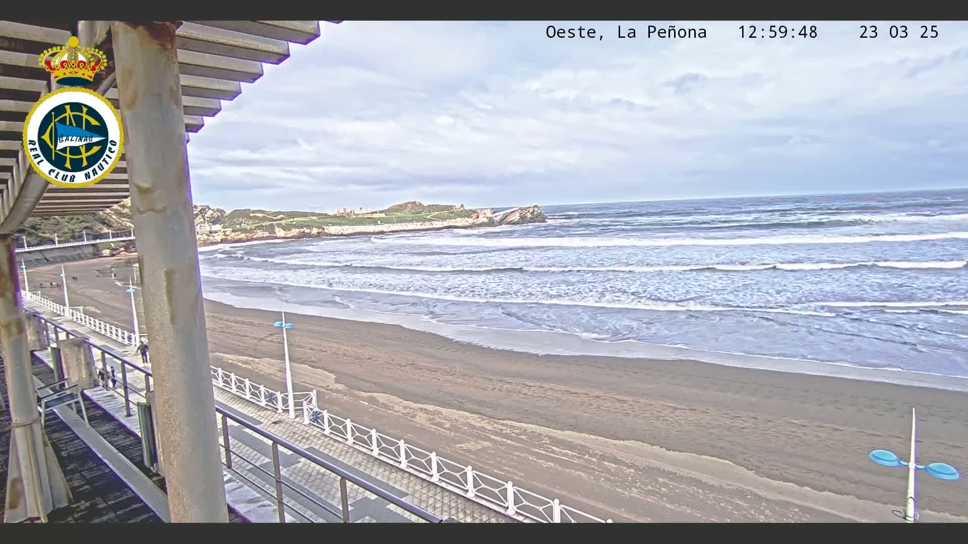 A sandy beach with gentle waves under a partly cloudy sky is viewed from an elevated vantage point, with a walkway and railing visible in the foreground.
