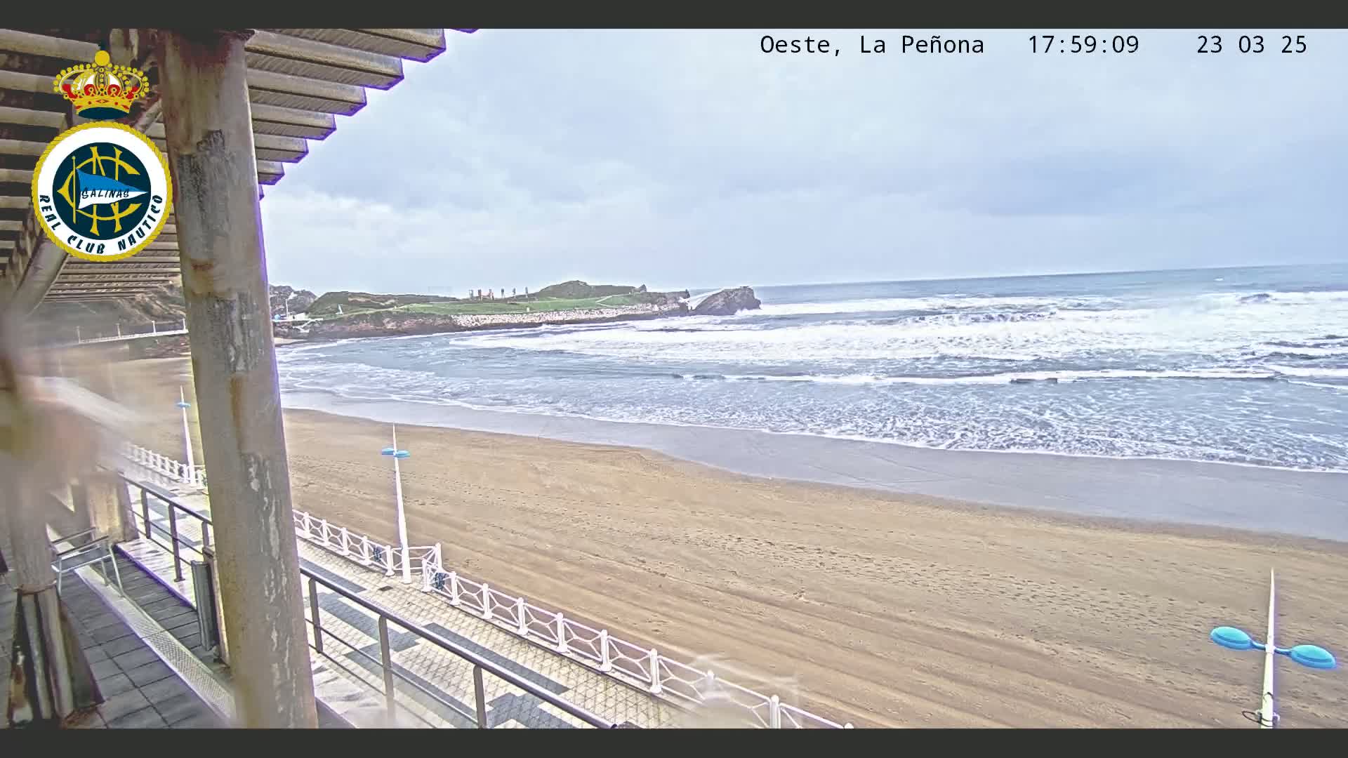 A sandy beach with moderate waves under an overcast sky is seen from a partially covered outdoor patio.