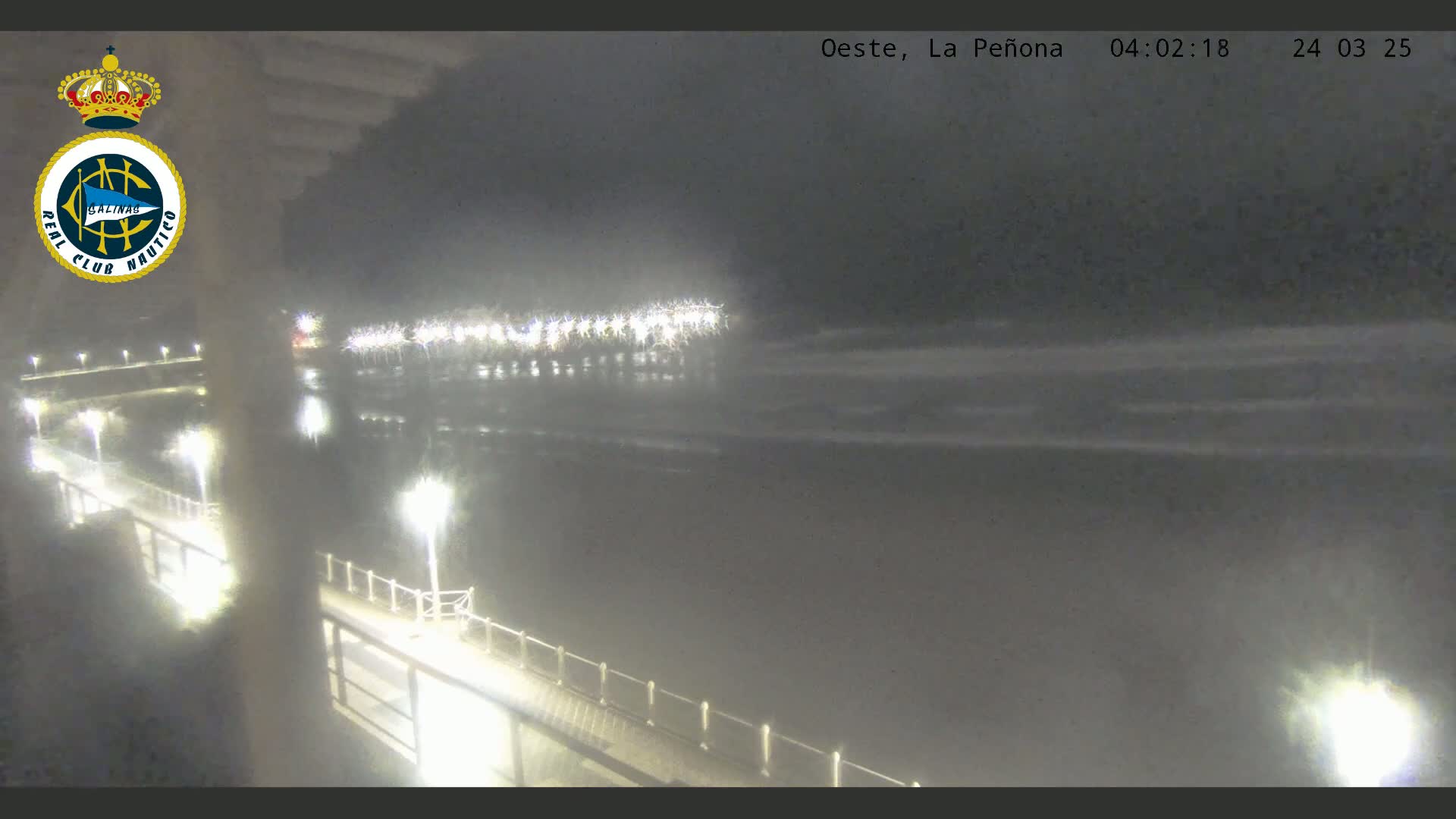 A nighttime view of a pier illuminated by lights, extending over dark water with gentle waves under a cloudy sky.