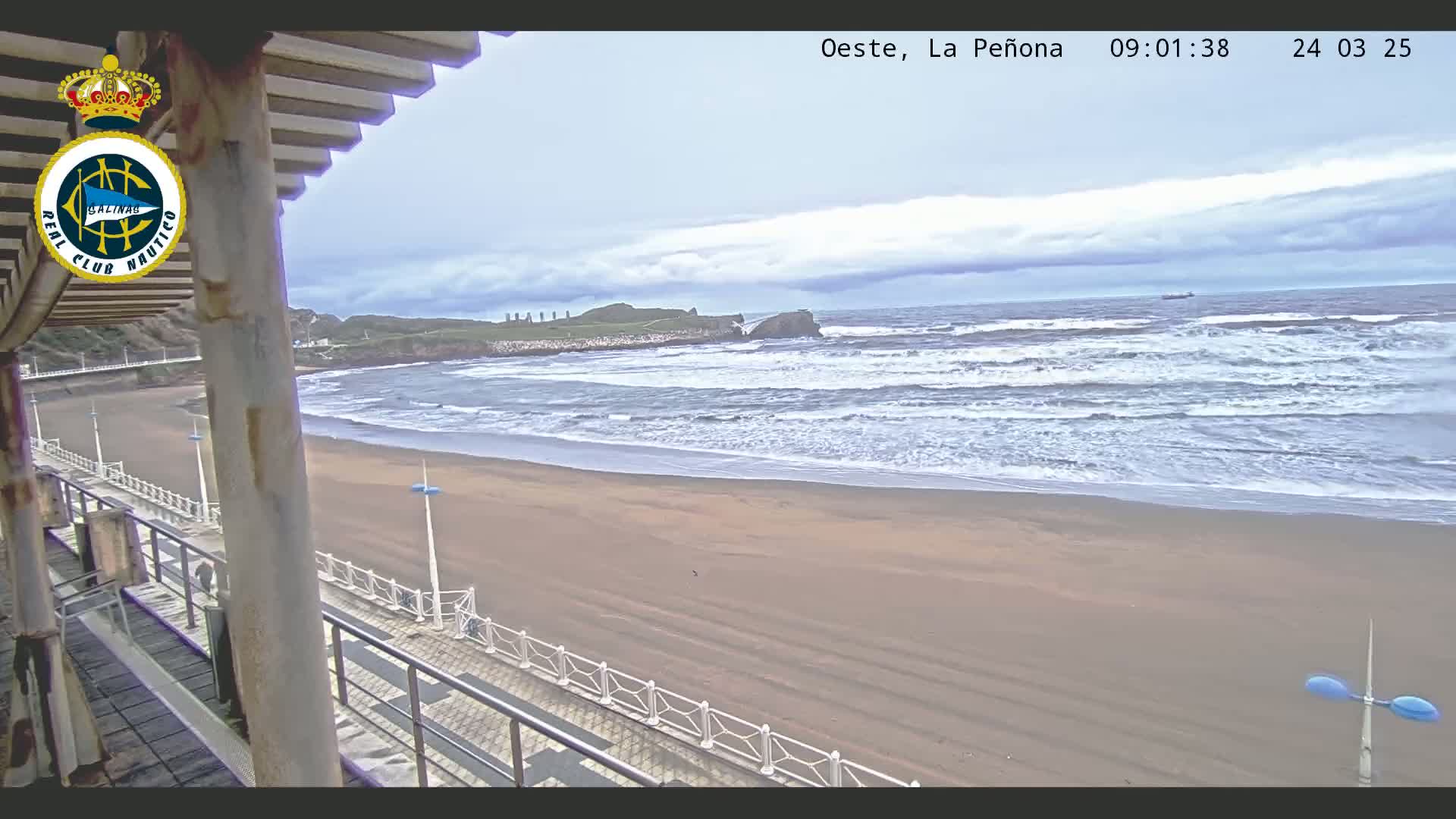 A sandy beach with moderate waves under a cloudy sky is viewed from a partially covered elevated structure.