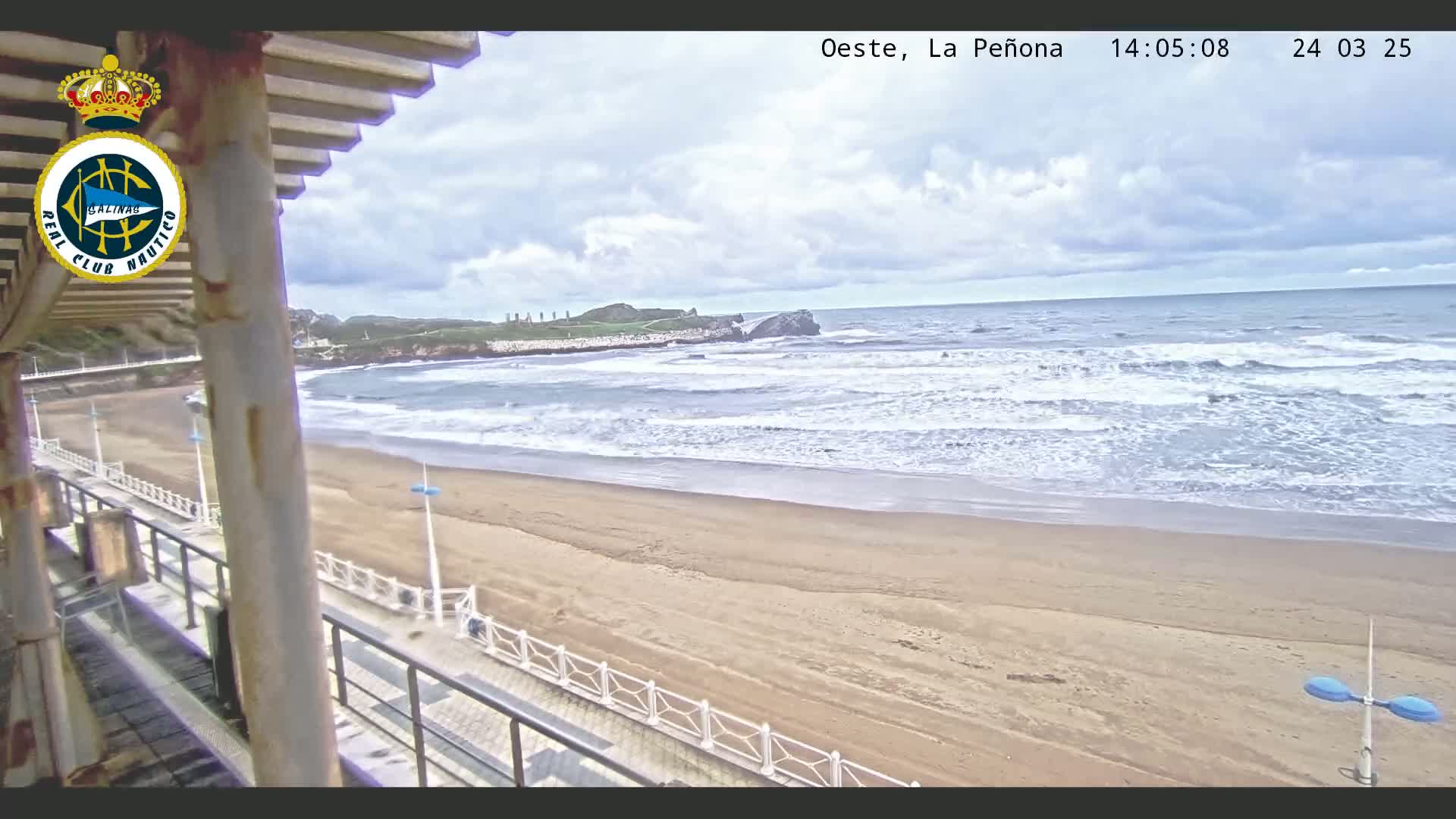 A sandy beach with moderate waves under a partly cloudy sky is viewed from a boardwalk with railings.