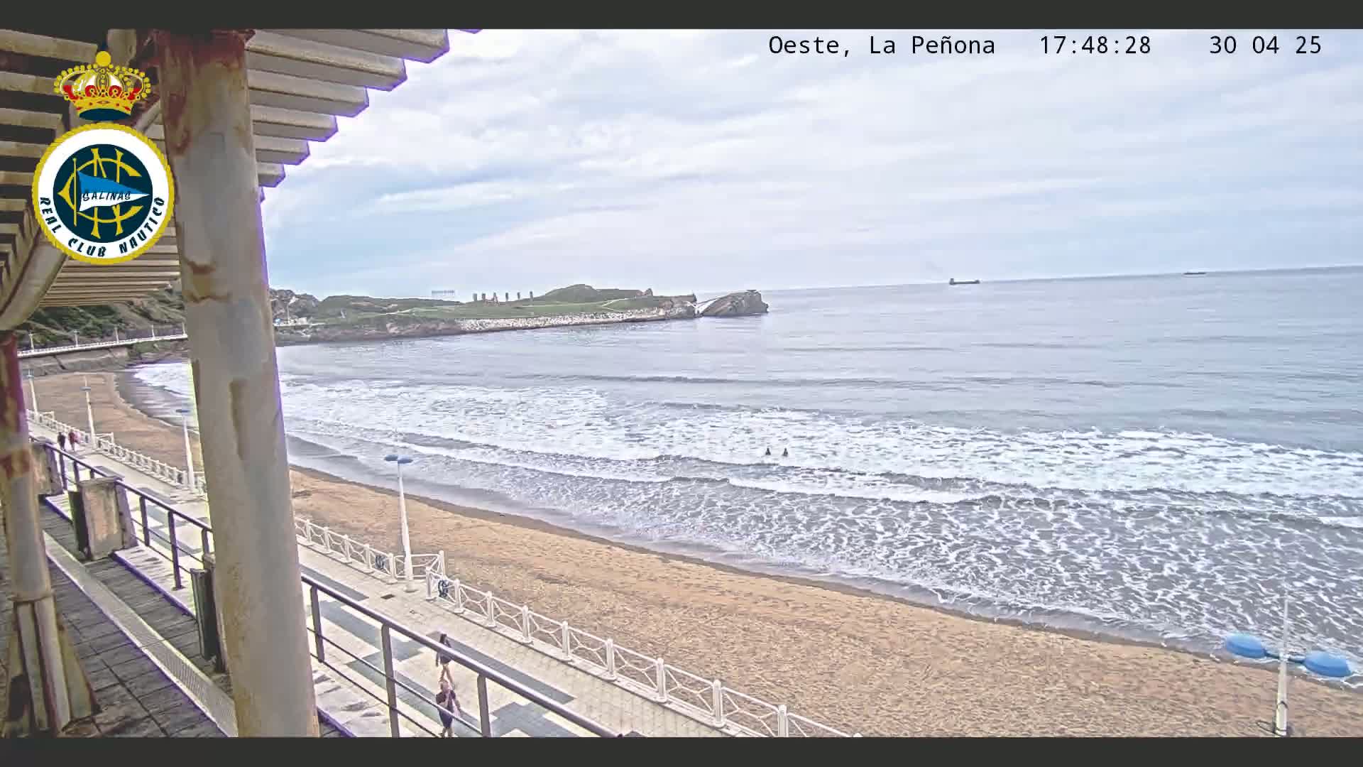 A sandy beach with gentle waves laps the shore under a partly cloudy sky, with a walkway and distant rocky outcropping visible.
