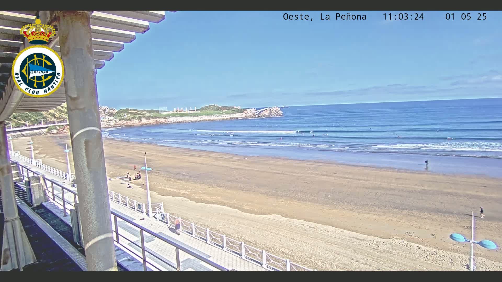 A sandy beach with several surfers in the ocean under a clear blue sky is viewed from an elevated position, overlooking a boardwalk.