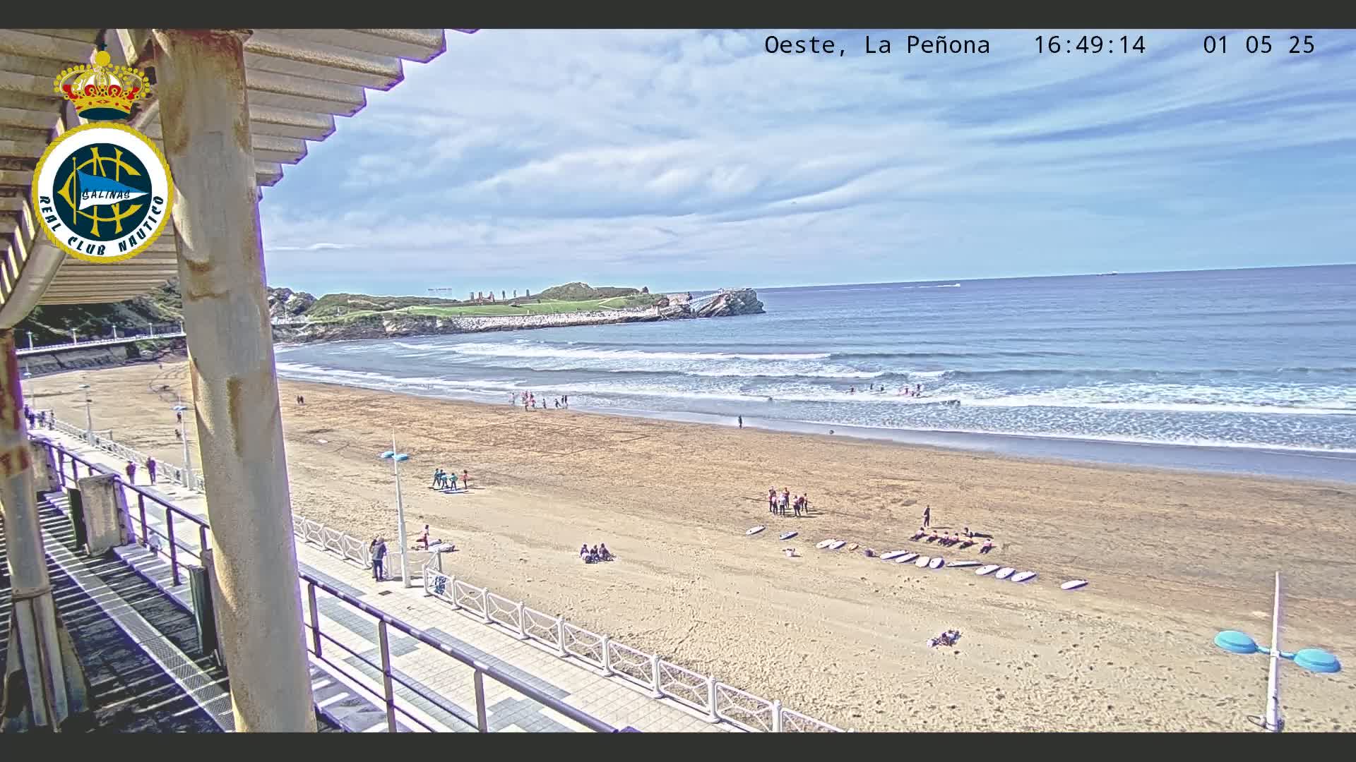 A sandy beach with gentle waves under a partly cloudy sky, viewed from an elevated position overlooking a boardwalk and a rocky outcropping in the distance.