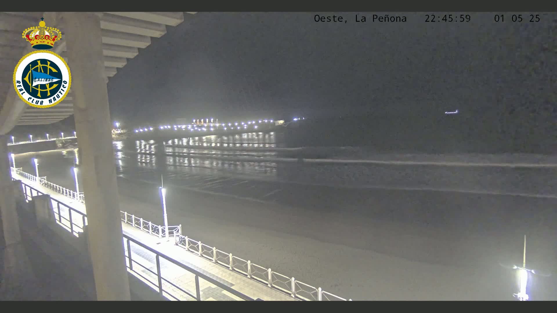 A nighttime view from a pier shows a calm ocean with gentle waves lapping at a sandy beach, illuminated by lights along the pier and a distant structure.