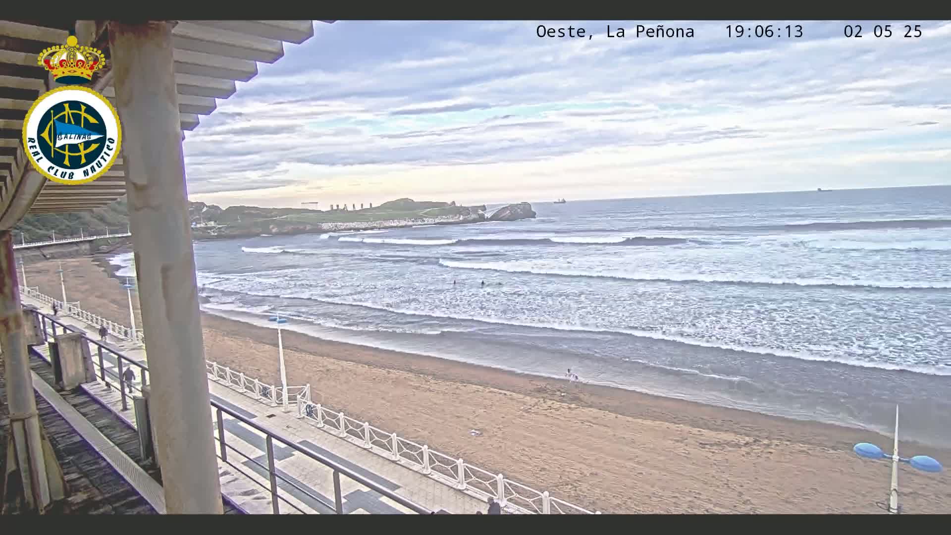 A sandy beach with gentle waves under a partly cloudy sky is seen from an elevated vantage point, with a walkway and a few people visible.