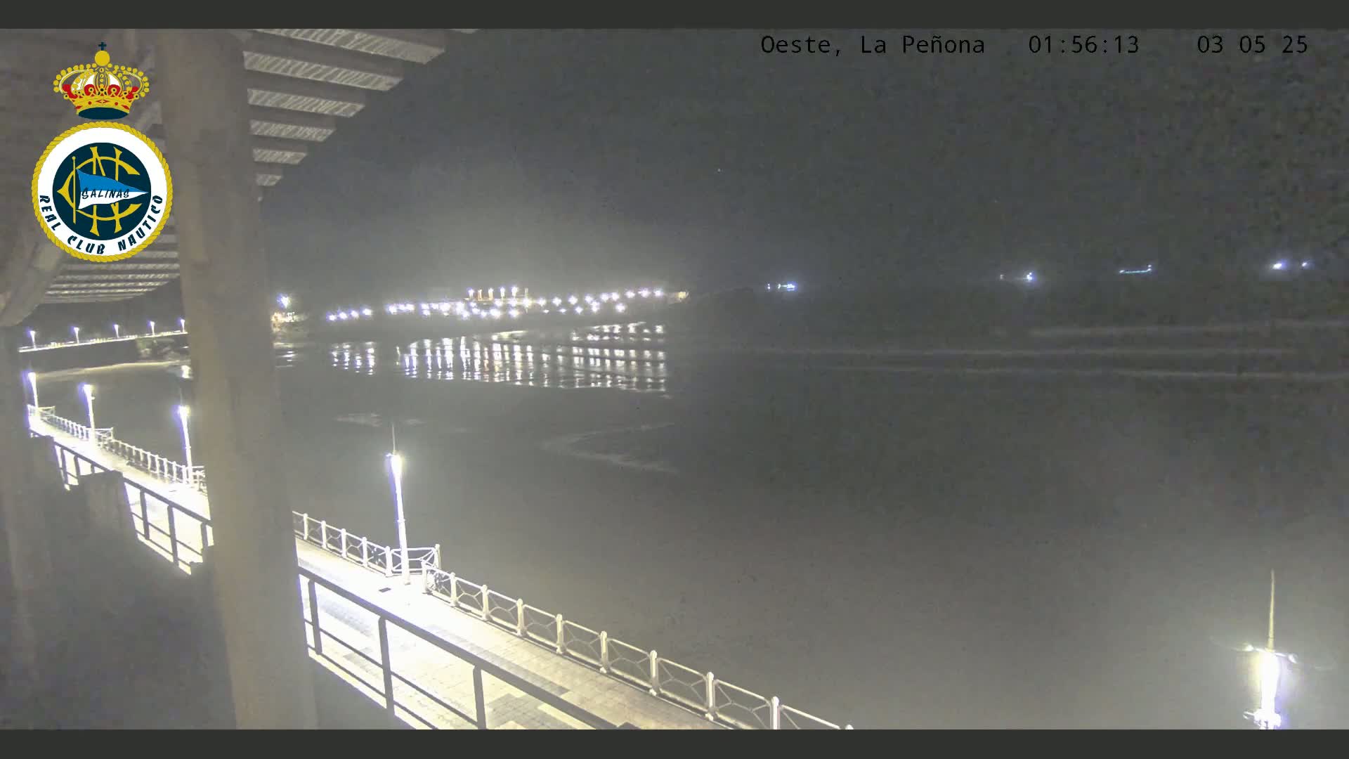 A nighttime view from a pier shows a calm body of water reflecting lights from a distant shore.
