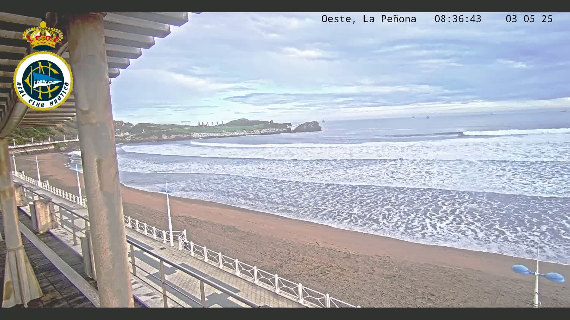 A sandy beach with gently rolling waves under a partly cloudy sky is viewed from an elevated position, showcasing a boardwalk and a distant rocky outcrop.