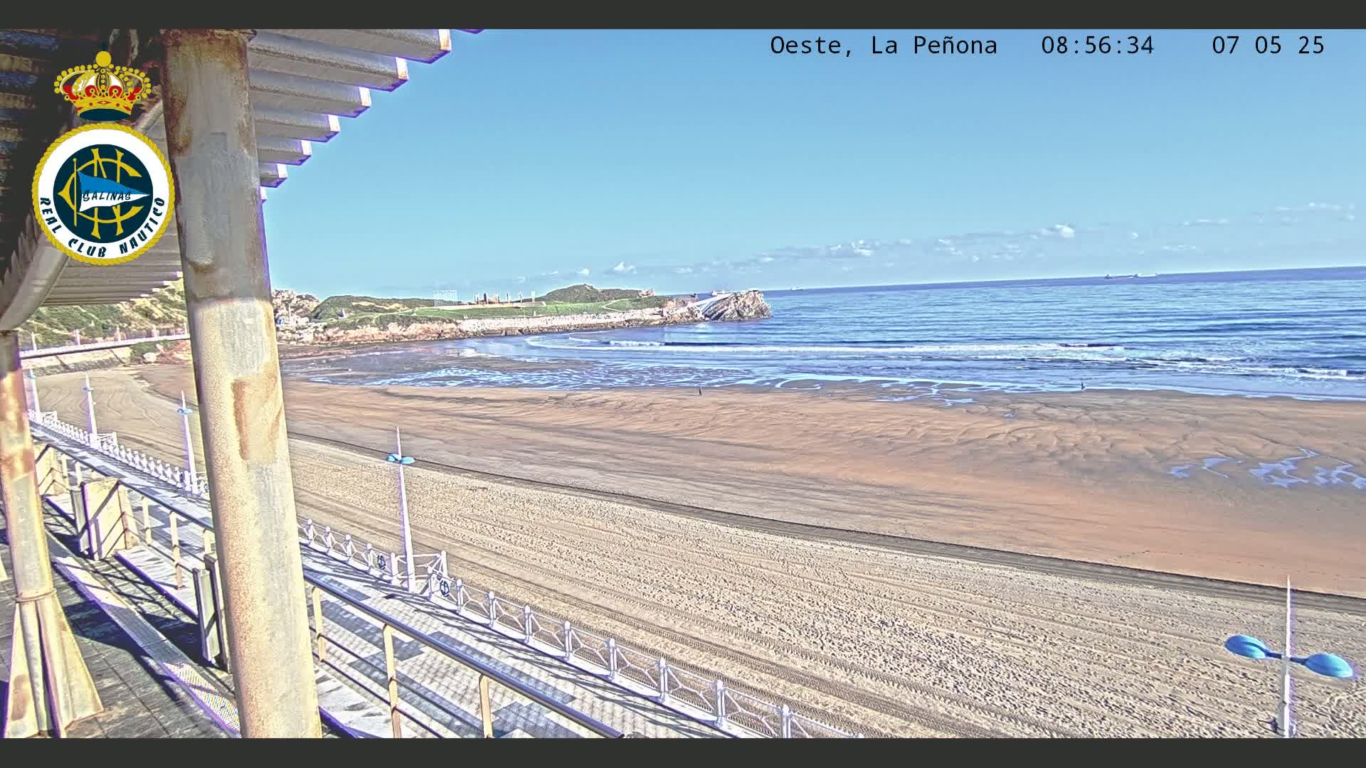 A sandy beach stretches to a calm ocean under a clear blue sky, viewed from an elevated vantage point with a walkway and railing in the foreground.