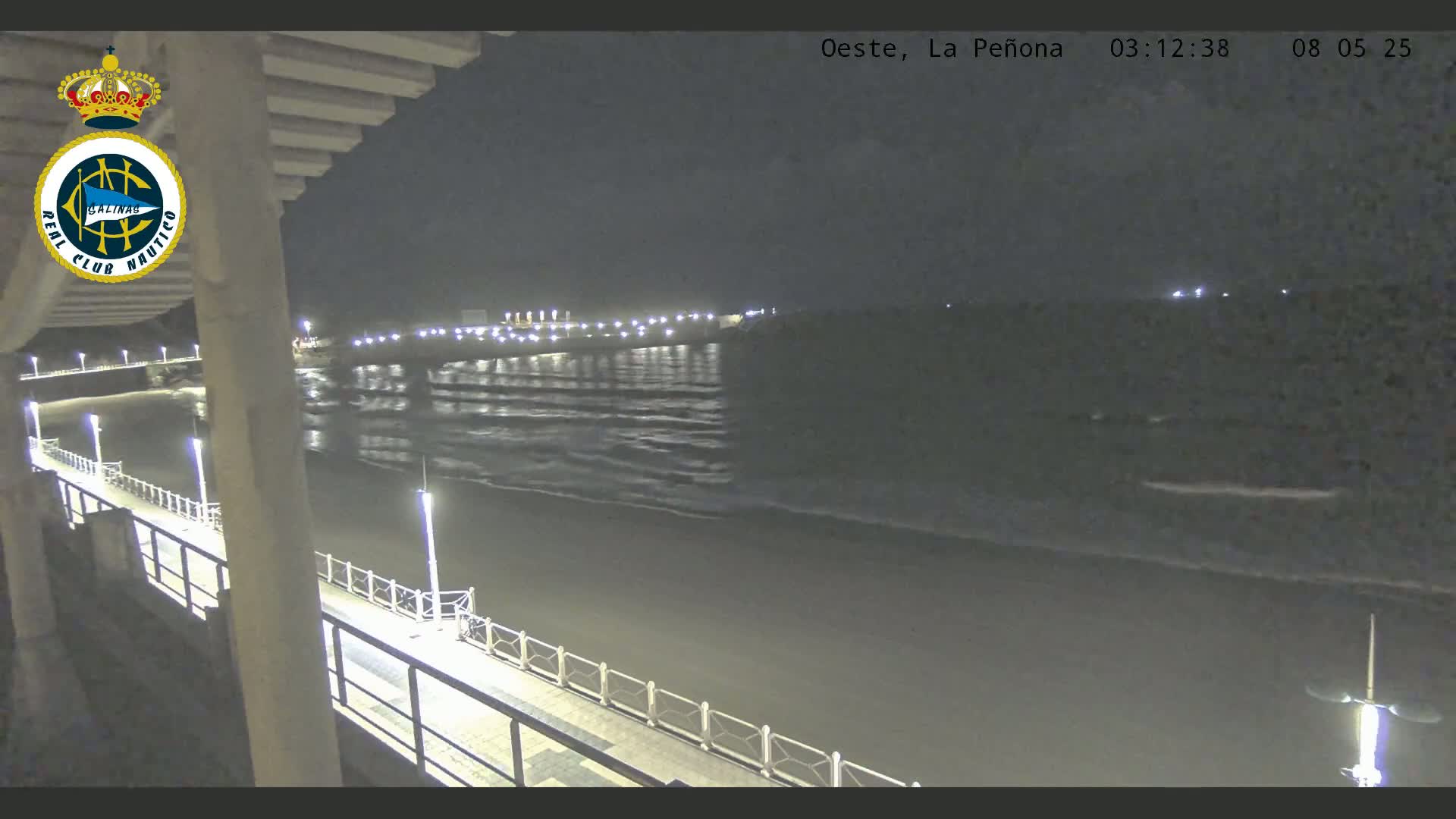 A nighttime view from a pier shows gently rolling waves on a dark beach under a cloudy sky.