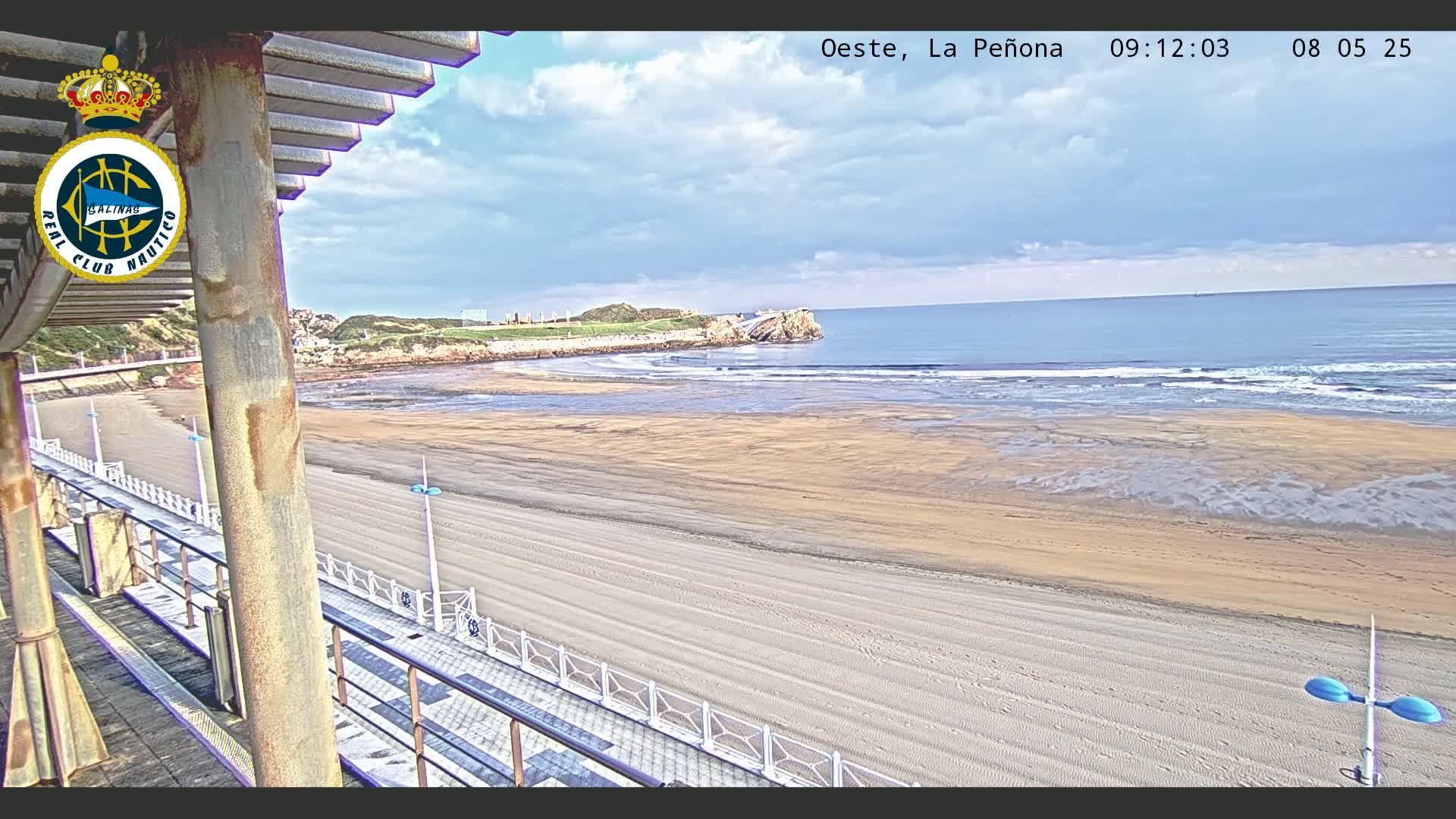 A sandy beach stretches out to a calm ocean under a partly cloudy sky, viewed from a raised vantage point with a walkway and railing in the foreground.