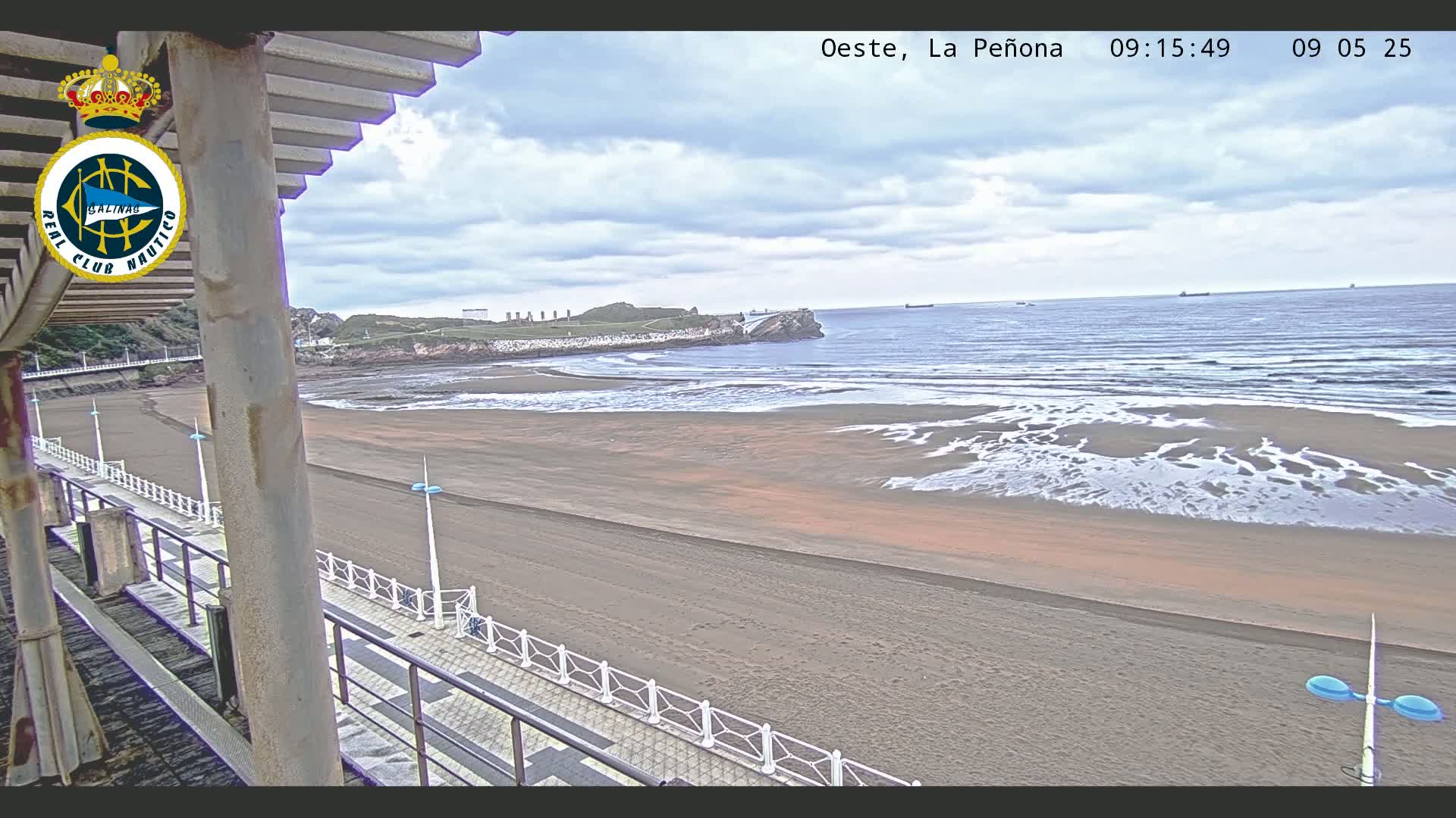 A sandy beach stretches out to a calm ocean under a cloudy sky, viewed from an elevated vantage point along a boardwalk.