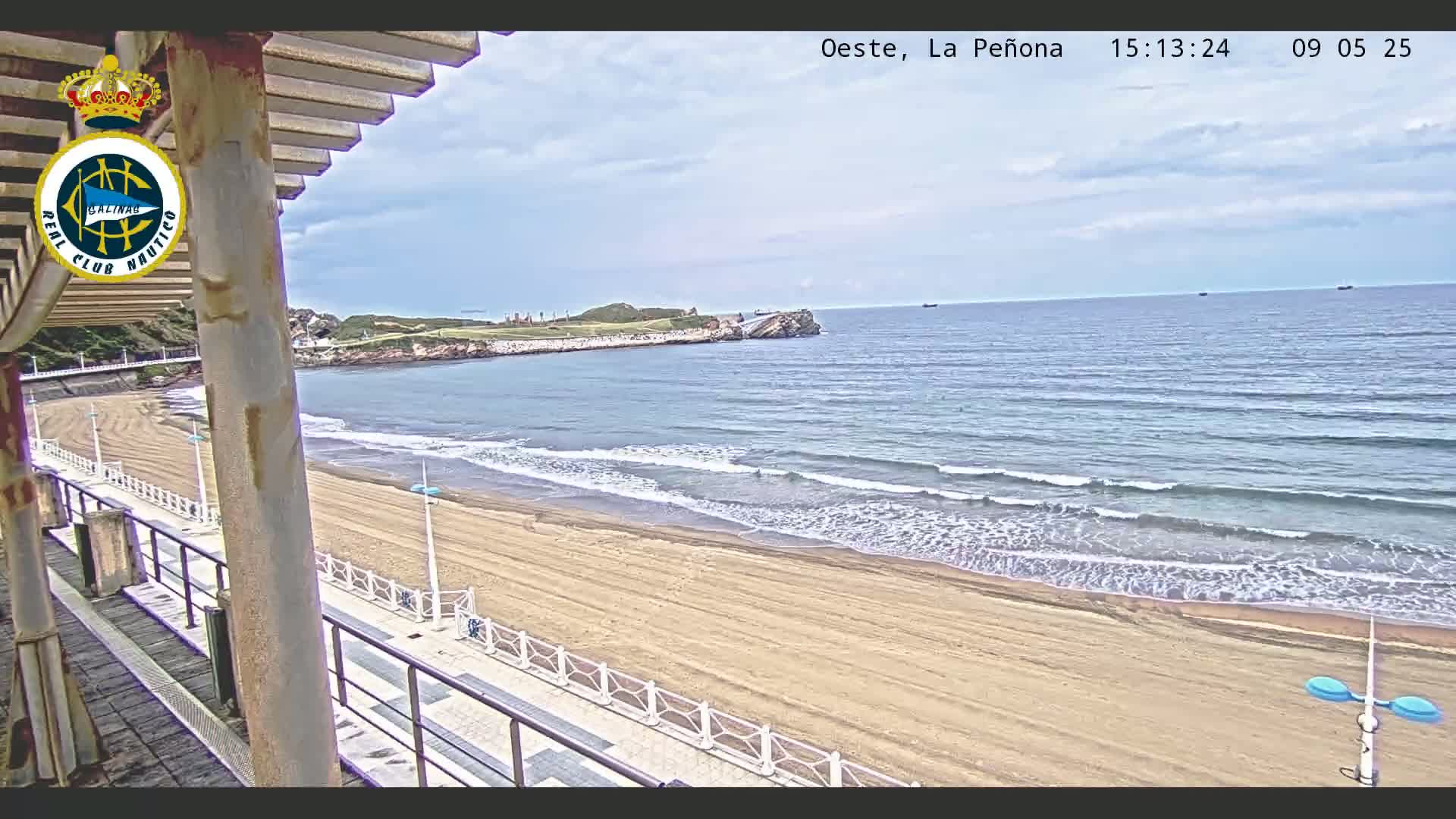 A sandy beach with gentle waves under a partly cloudy sky is viewed from a covered boardwalk.