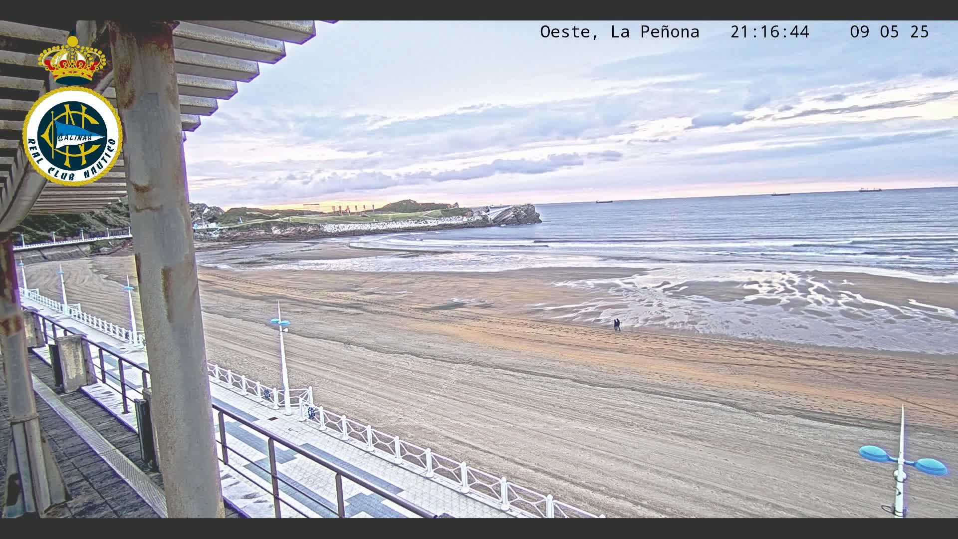 A sandy beach stretches to a calm ocean under a cloudy, twilight sky, with a paved walkway and railing visible in the foreground.