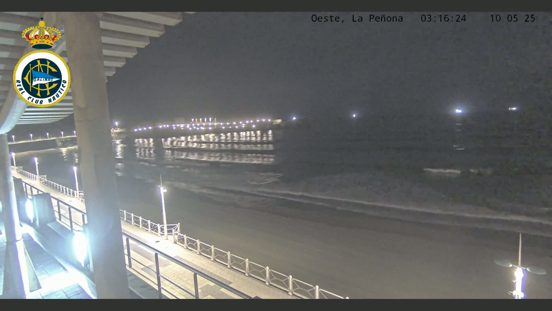 A nighttime view from an elevated position shows a dark beach with gentle waves lapping the shore, a lit walkway, and a body of water with lights reflecting on its surface in the distance.