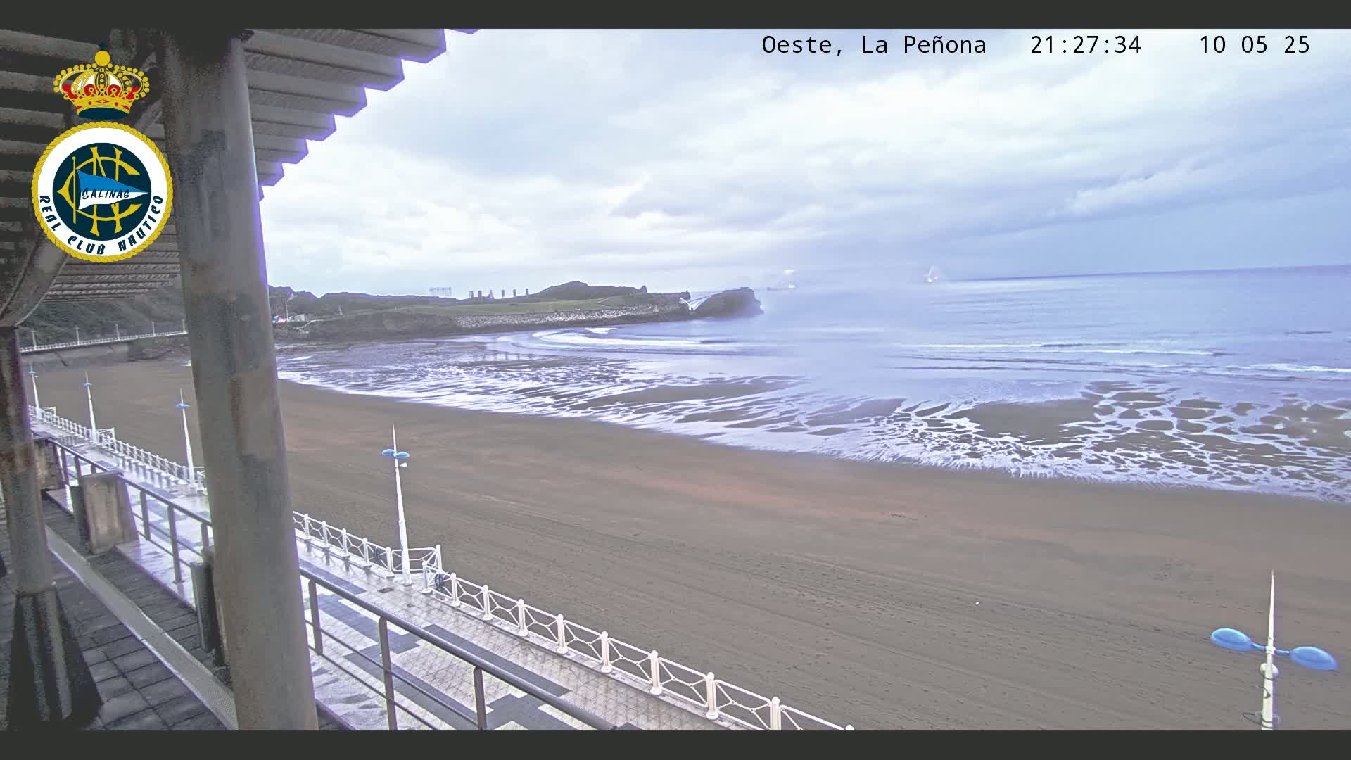 A mostly overcast day shows a sandy beach with gentle waves lapping the shore, viewed from an elevated position near a walkway with railings.