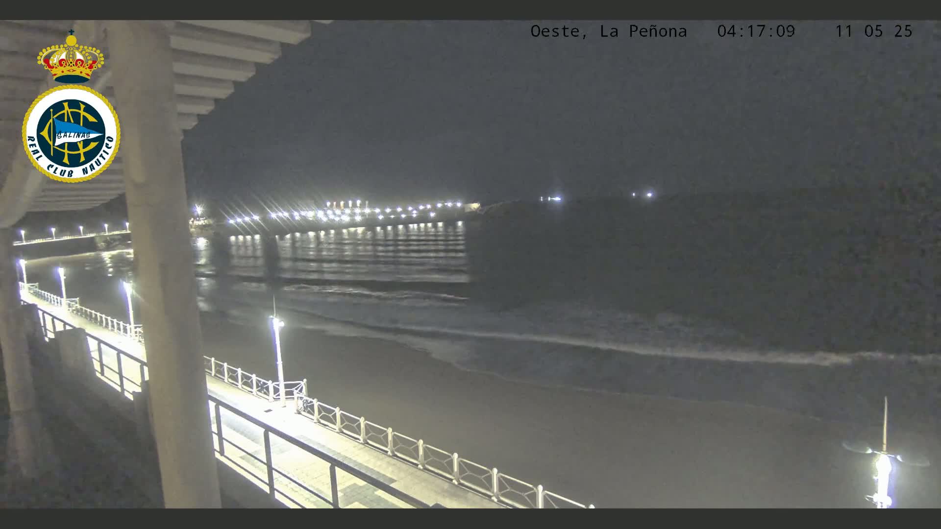 A nighttime view from a pier shows a dark beach with gently lapping waves and a brightly lit shoreline in the distance.
