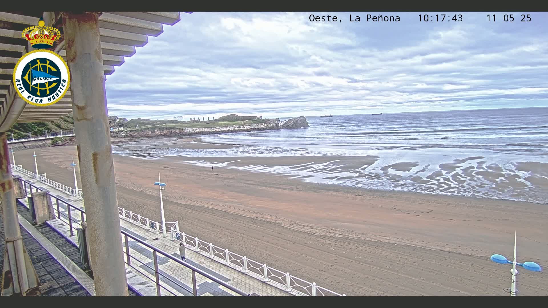 A sandy beach with gentle waves under a mostly cloudy sky is seen from a seaside boardwalk.
