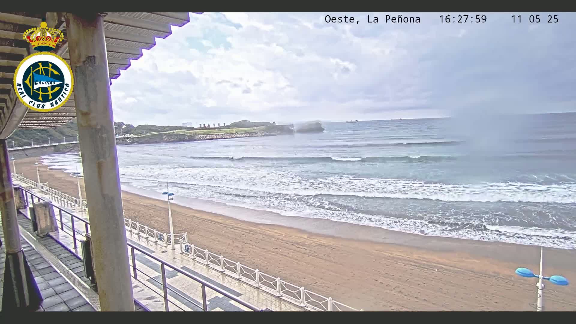 A sandy beach with gentle waves under a cloudy sky is viewed from a balcony overlooking a boardwalk.
