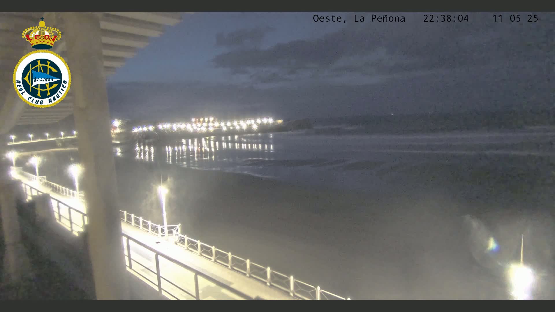 A nighttime view from a pier shows a calm body of water reflecting lights from a distant shoreline under a partly cloudy sky.