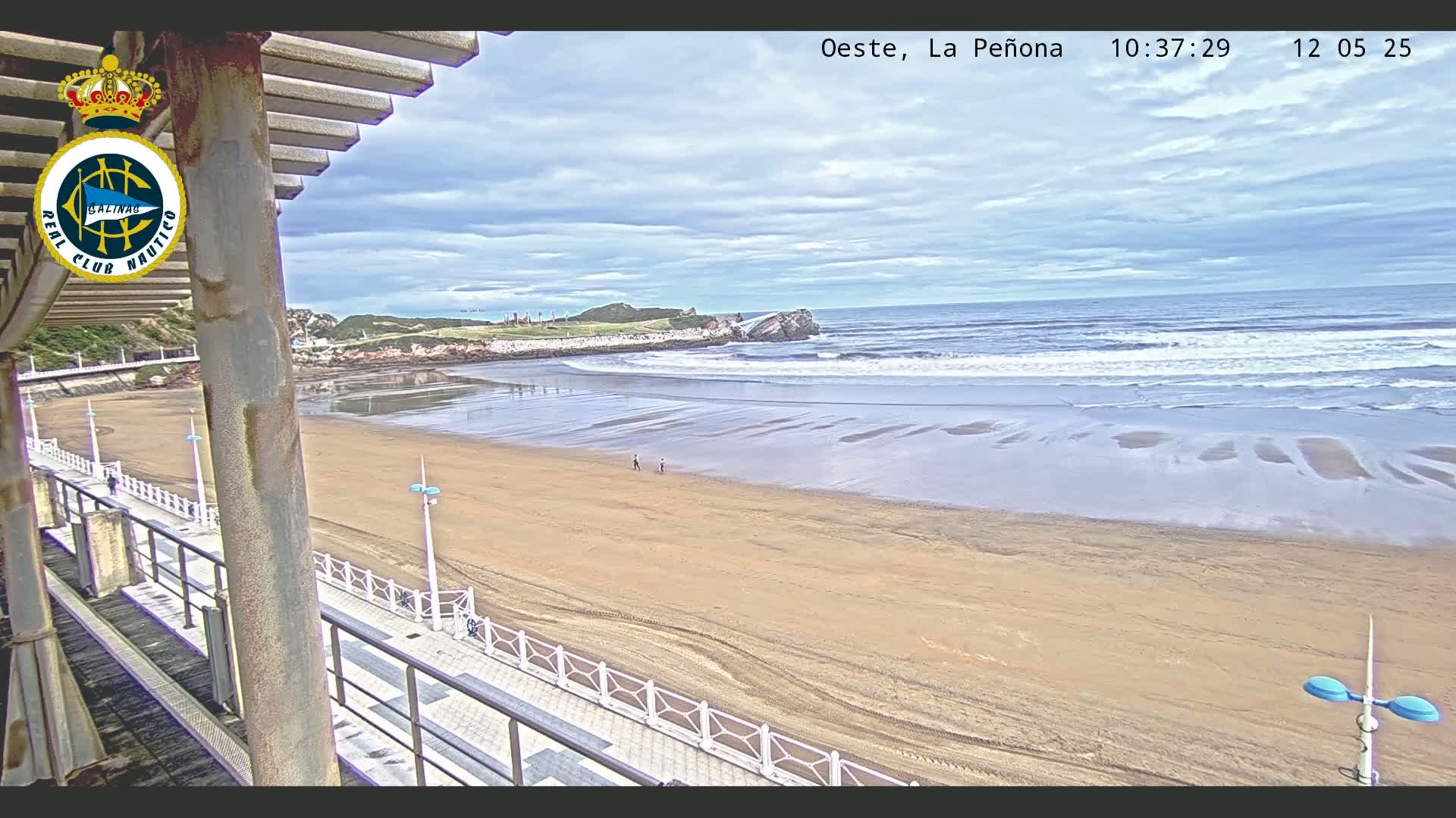 A sandy beach with gentle waves under a partly cloudy sky is viewed from an elevated position, showing a boardwalk and a few people walking along the shore.