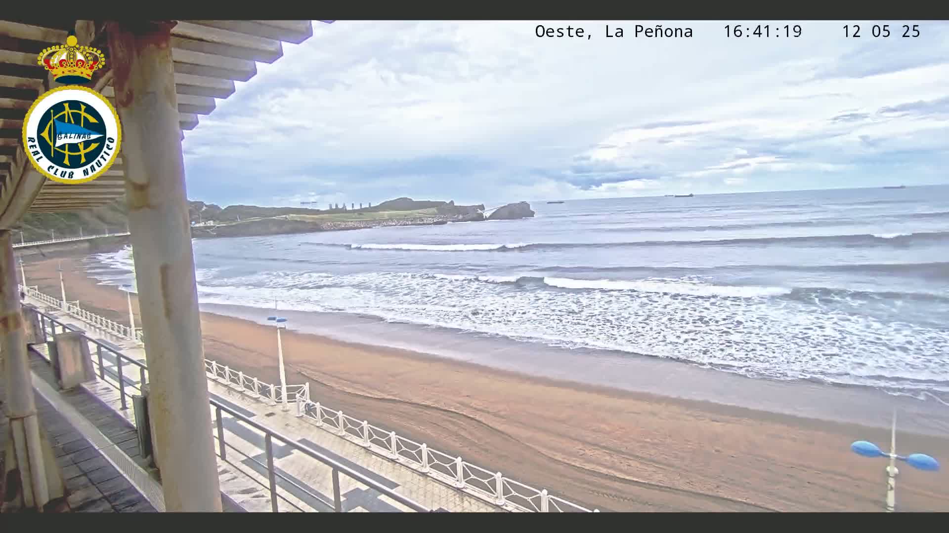A sandy beach with gentle waves under a partly cloudy sky is seen from a high vantage point, overlooking a boardwalk with railings.