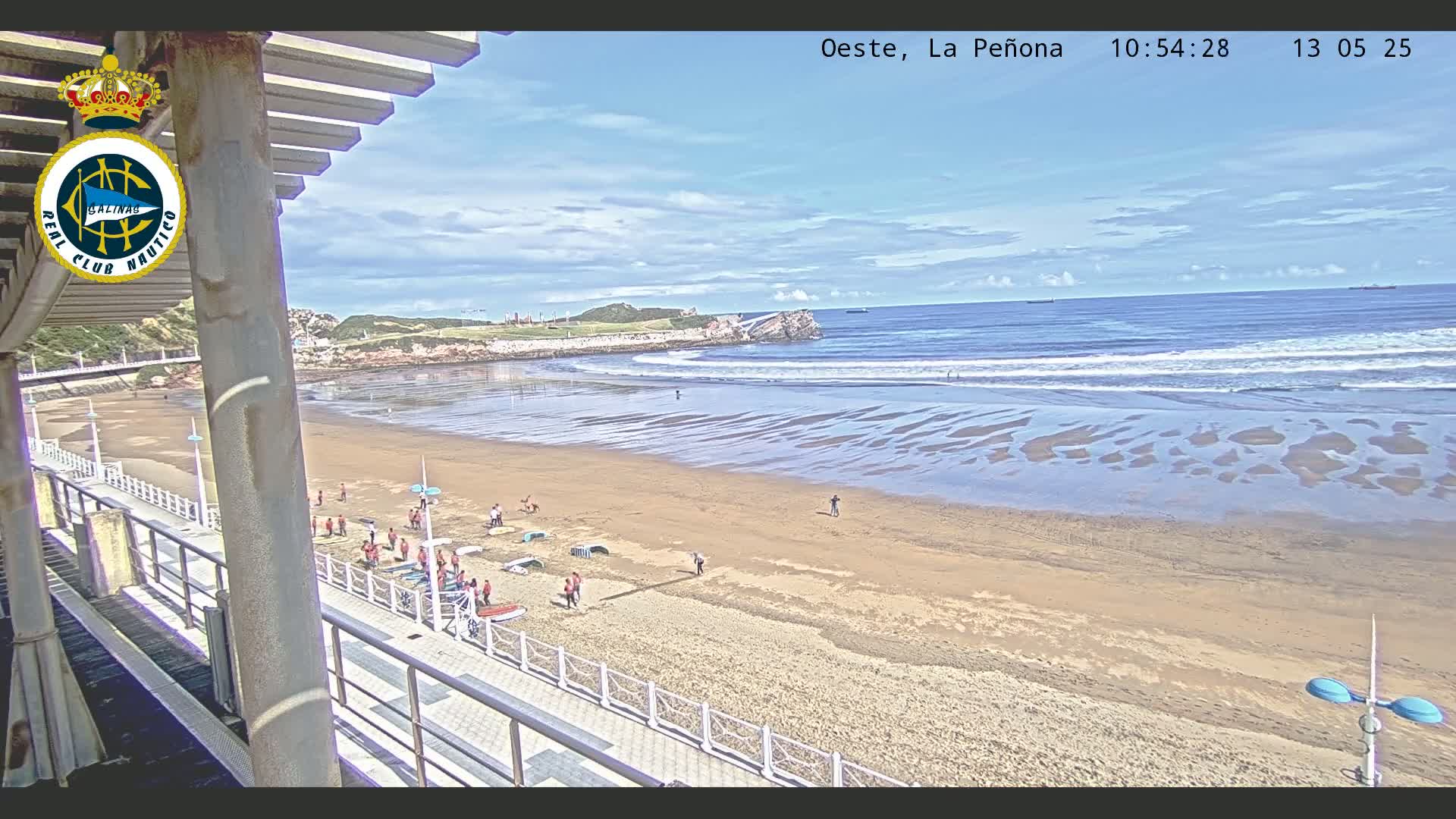 A sandy beach with a few people on it,  calm ocean waves, and a partly cloudy sky is seen from an elevated vantage point.