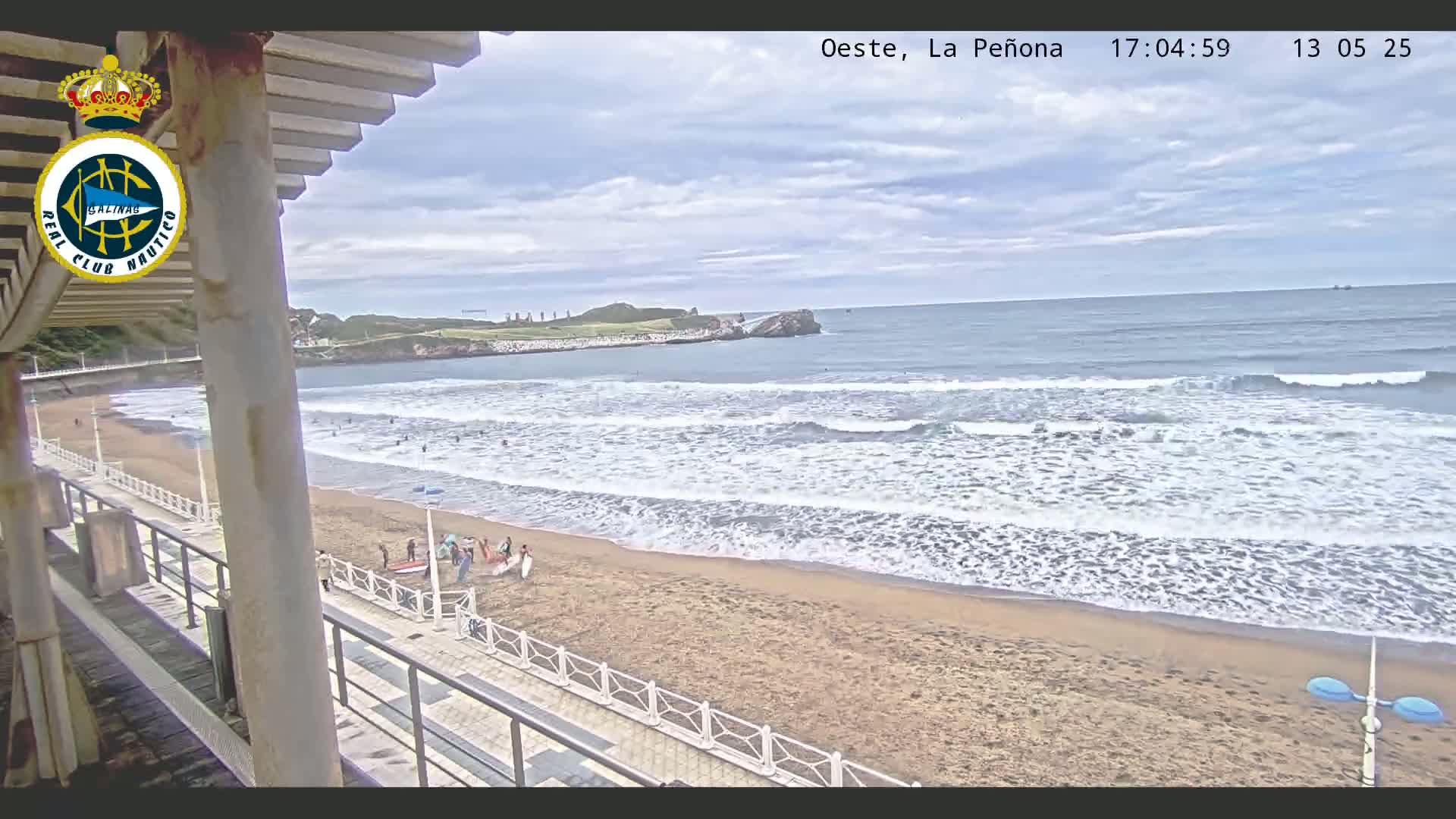 A sandy beach with gentle waves under a partly cloudy sky is viewed from a structure overlooking the ocean, with several people visible on the beach.