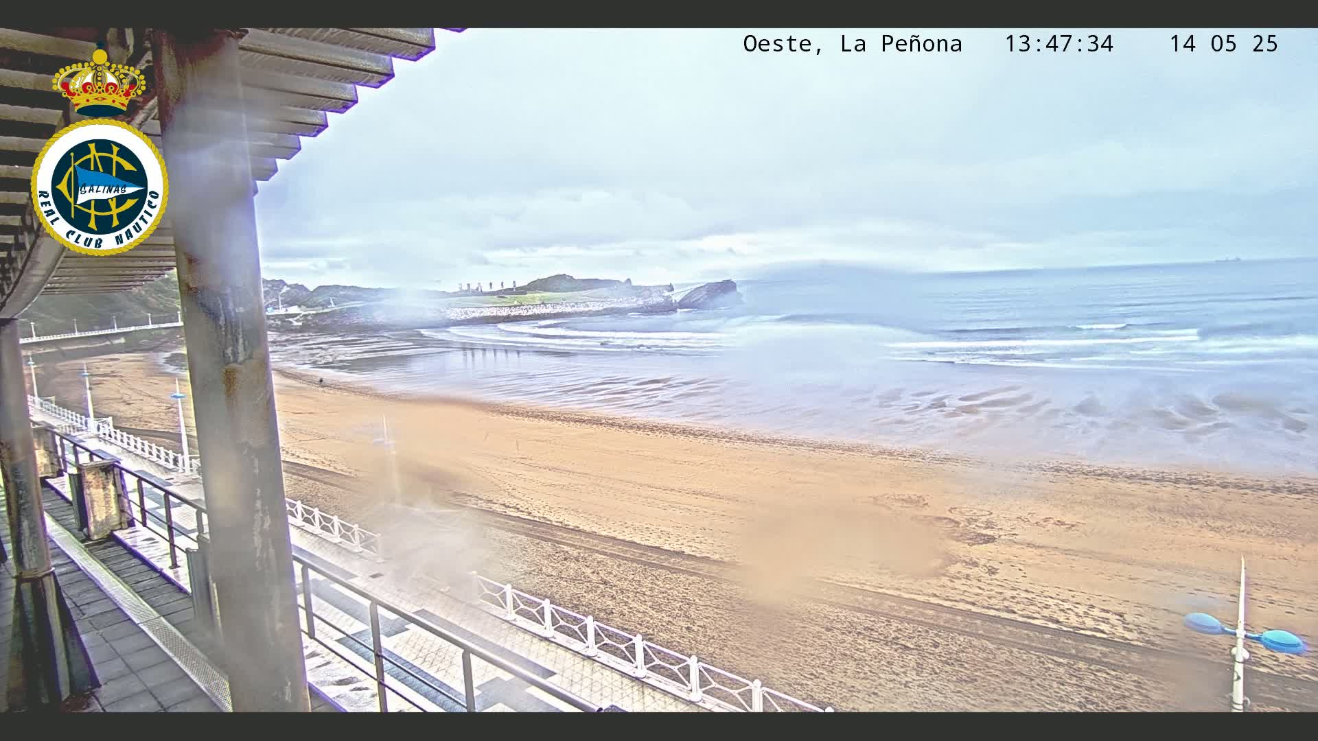 A hazy view from a balcony shows a sandy beach with gentle waves lapping the shore under a cloudy sky.