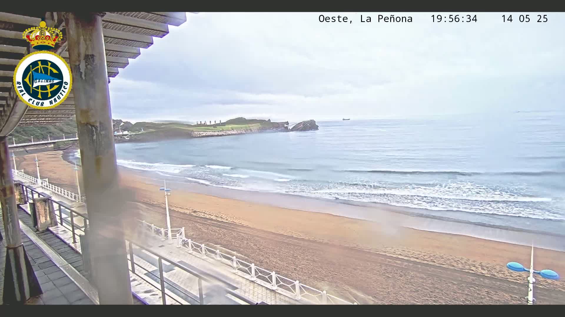 An overcast day reveals a sandy beach with gentle waves lapping the shore, a distant rocky outcrop, and a walkway visible from a partially covered outdoor vantage point.