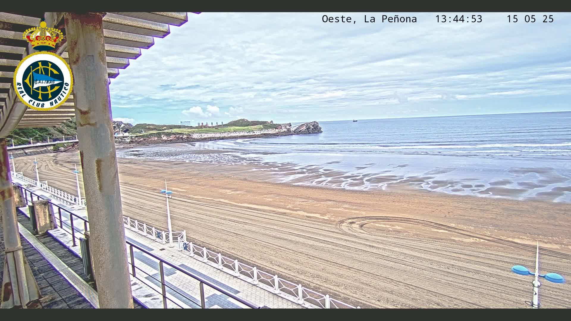 A sandy beach with a calm ocean under a partly cloudy sky is viewed from a boardwalk with a railing.