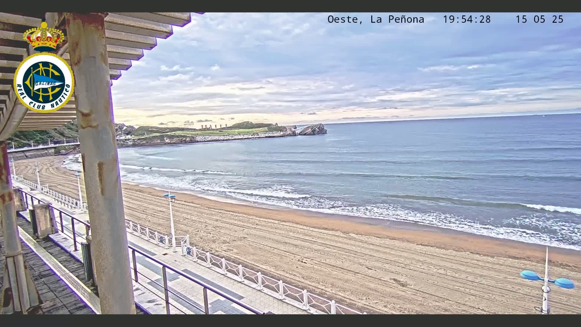 A sandy beach with gentle waves under a partly cloudy sky is viewed from a seaside structure.