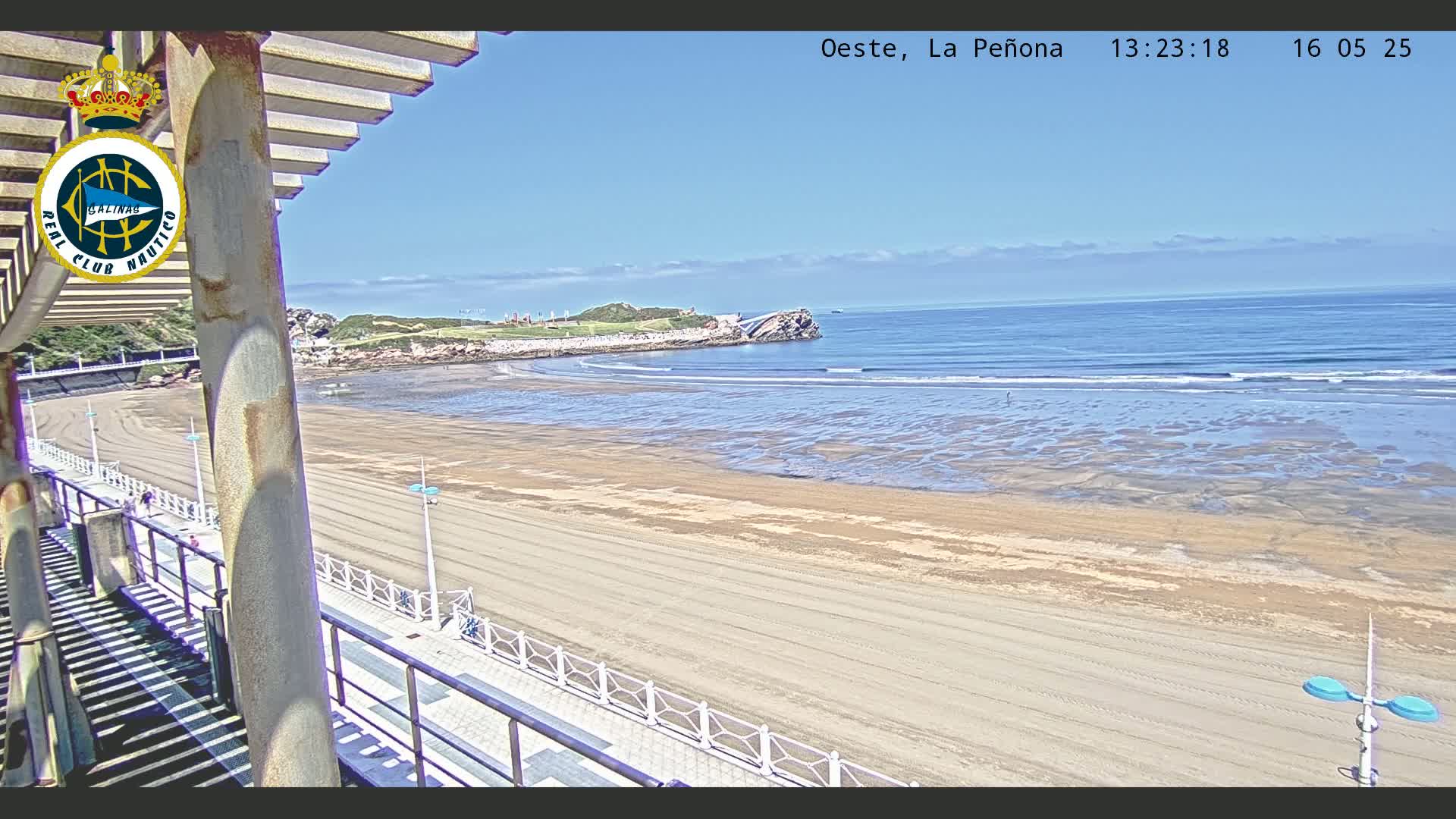 A sandy beach stretches out to a calm ocean under a clear blue sky, viewed from an elevated vantage point with a railing and columns in the foreground.