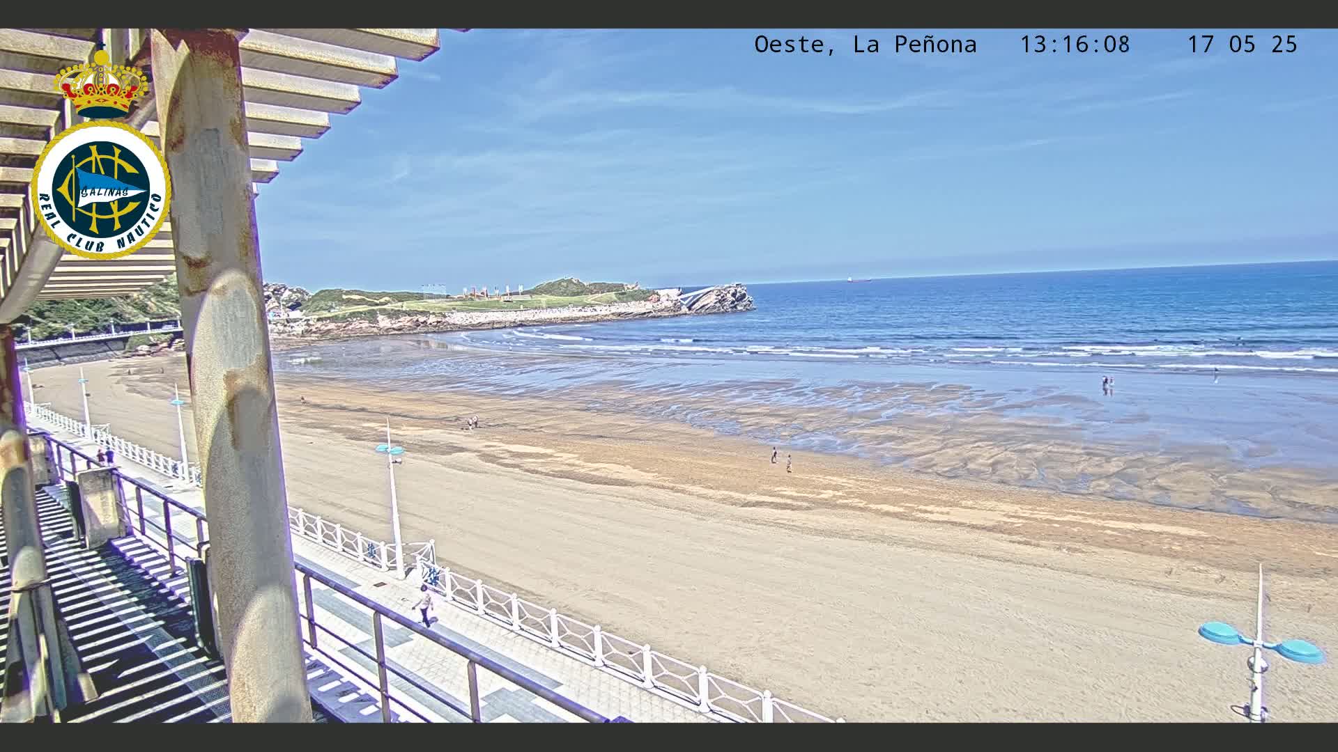 A sandy beach with a few people on it stretches toward a calm ocean under a clear blue sky, viewed from an elevated vantage point with a railing and shaded structure.