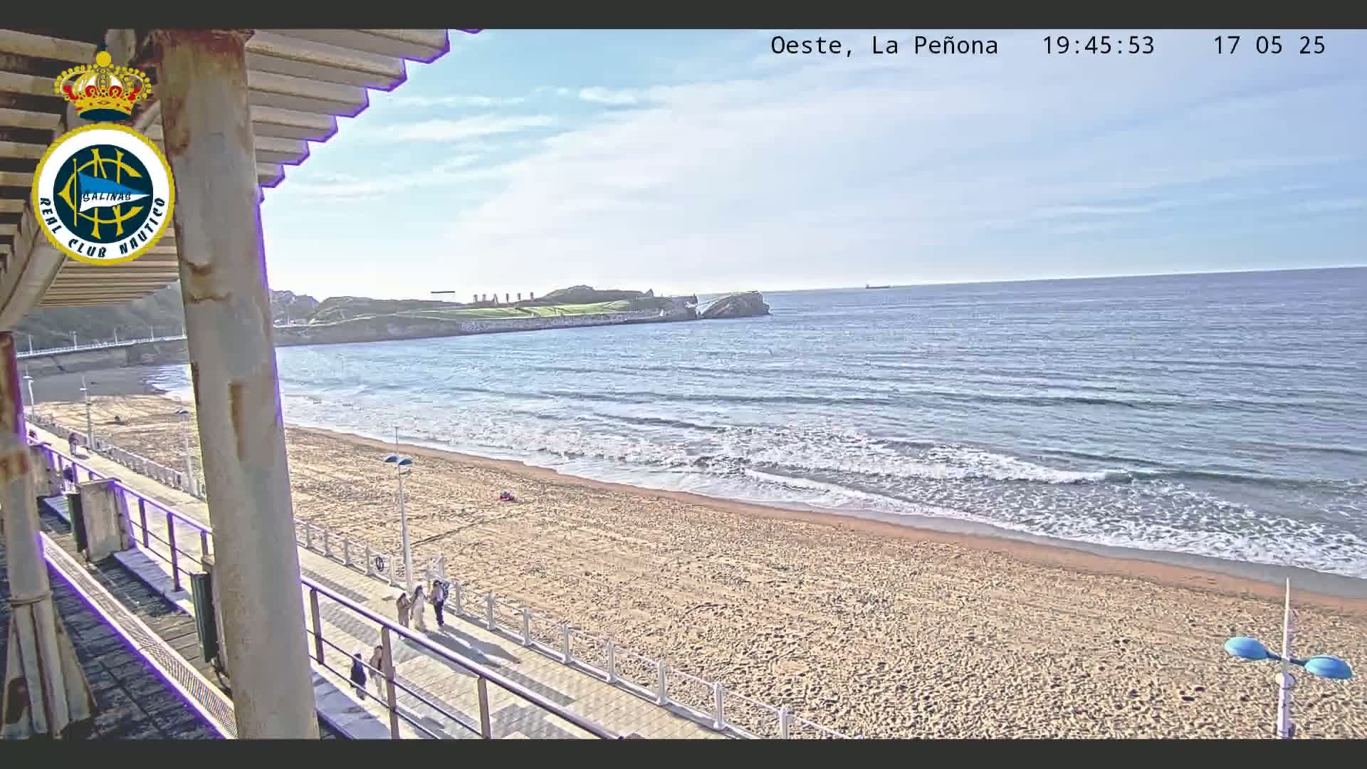 A sandy beach with calm waves and a few people walking along a boardwalk, under a partly cloudy sky.