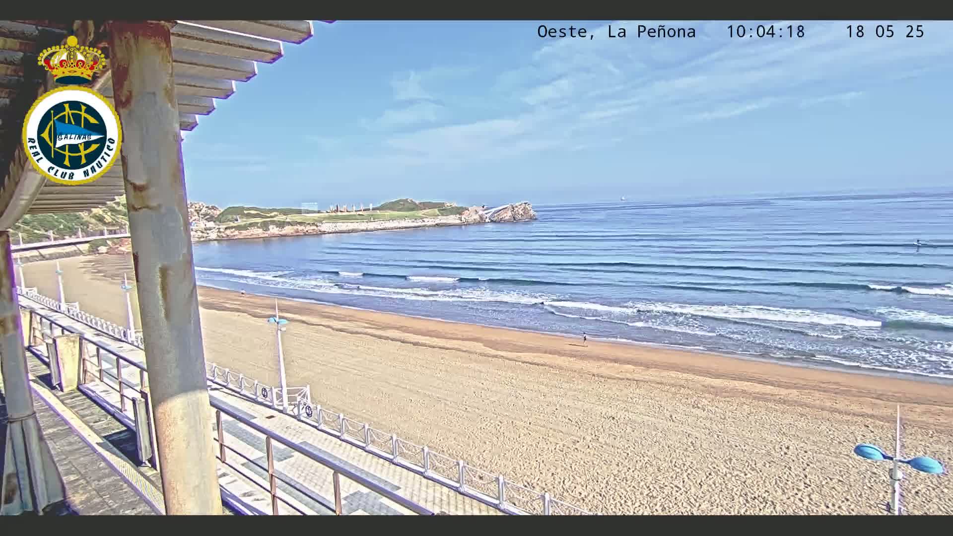 A sandy beach with gentle waves under a mostly clear sky is viewed from a high vantage point, showing a boardwalk and a rocky outcropping in the distance.