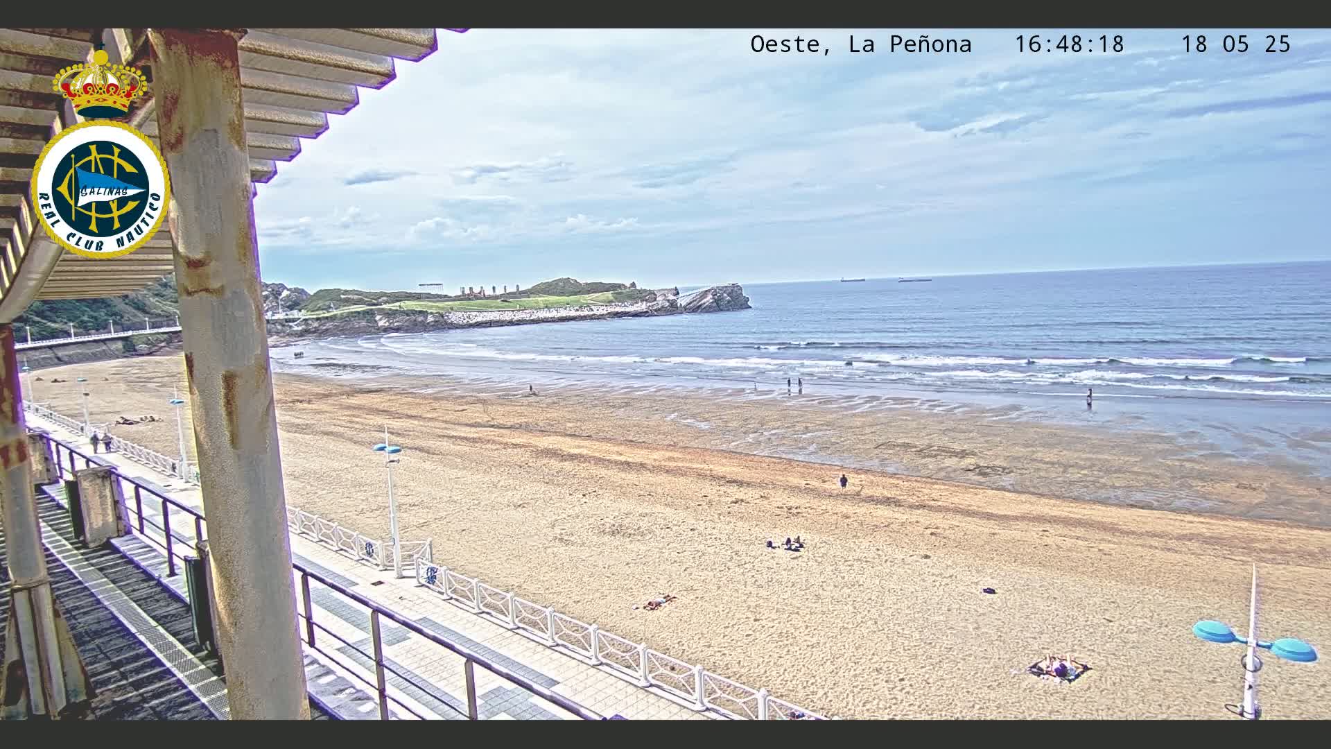 A mostly sunny day shows a long sandy beach with a few people scattered on it, a calm ocean with small waves, and a rocky headland in the distance.