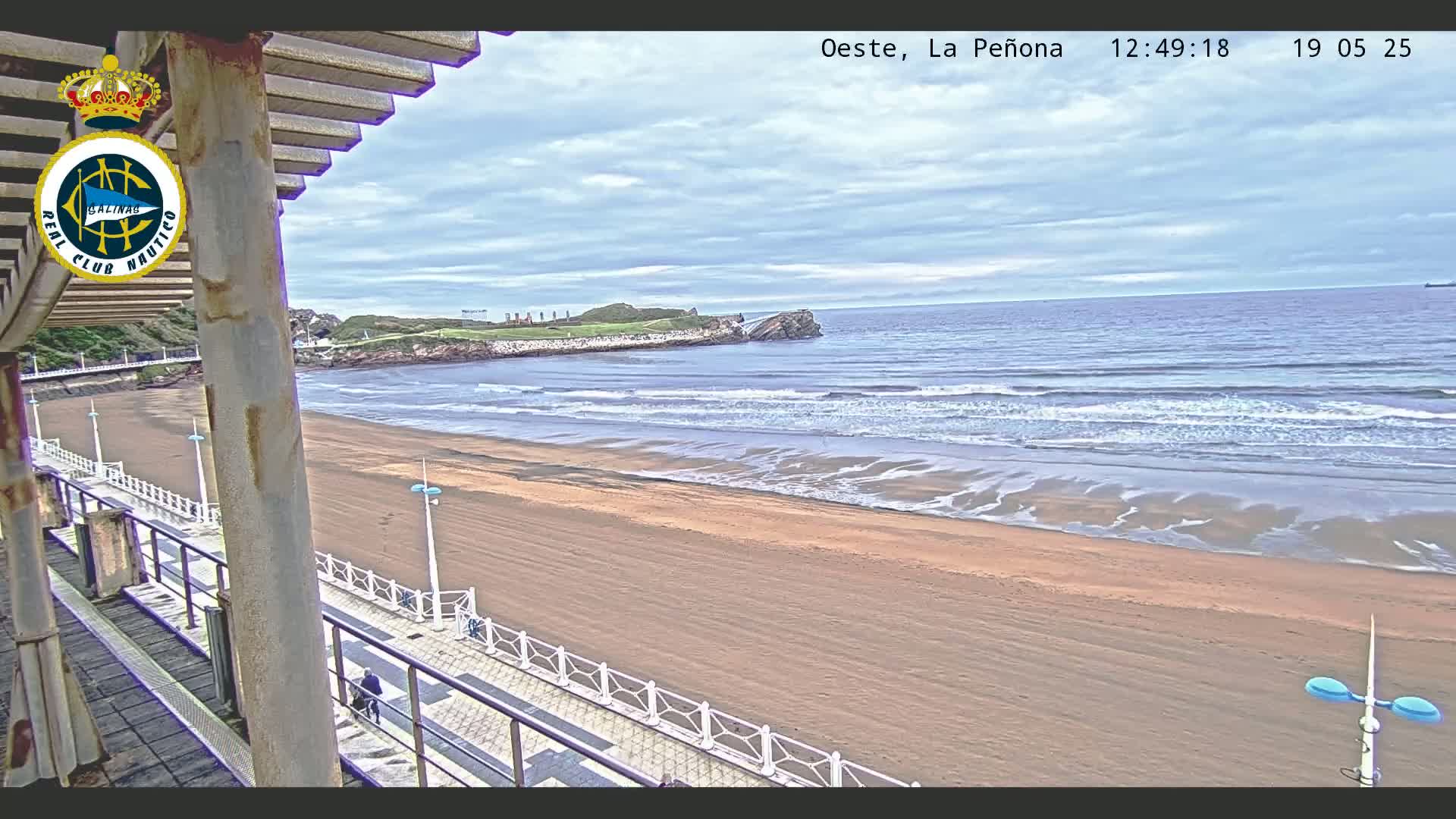 A sandy beach with gentle waves under a cloudy sky is seen from an elevated vantage point, overlooking a paved walkway and a structure with a slatted roof.