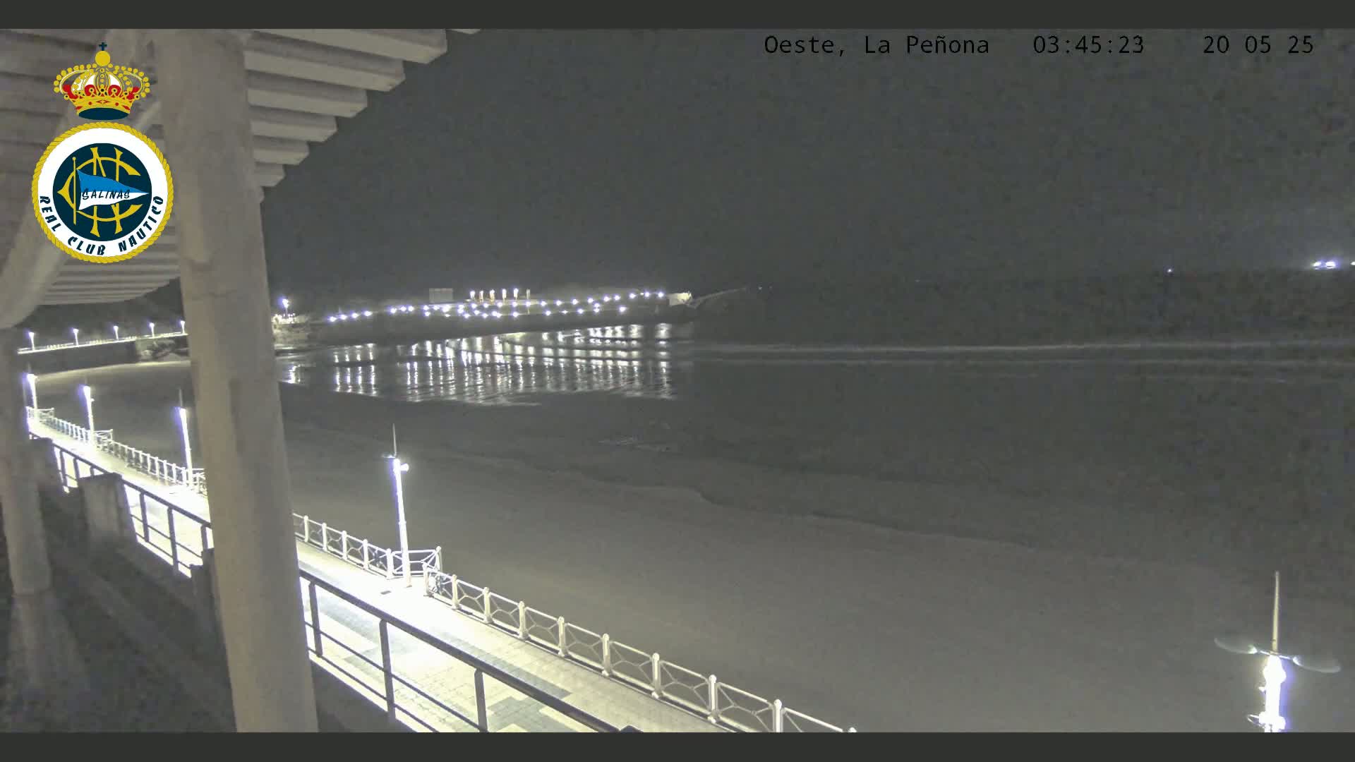 A nighttime view from an elevated position shows a dimly lit beach, a pier with lights, and calm ocean water reflecting the lights.