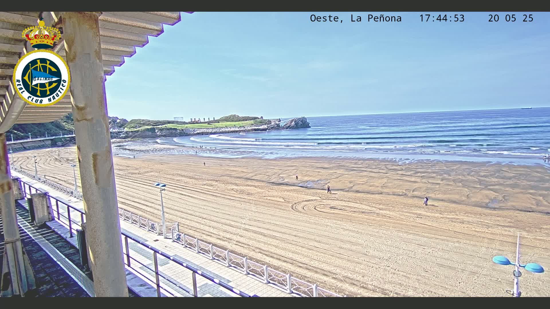 A sandy beach with gentle waves under a clear blue sky is viewed from an elevated position, showing a boardwalk and a few people walking on the sand.