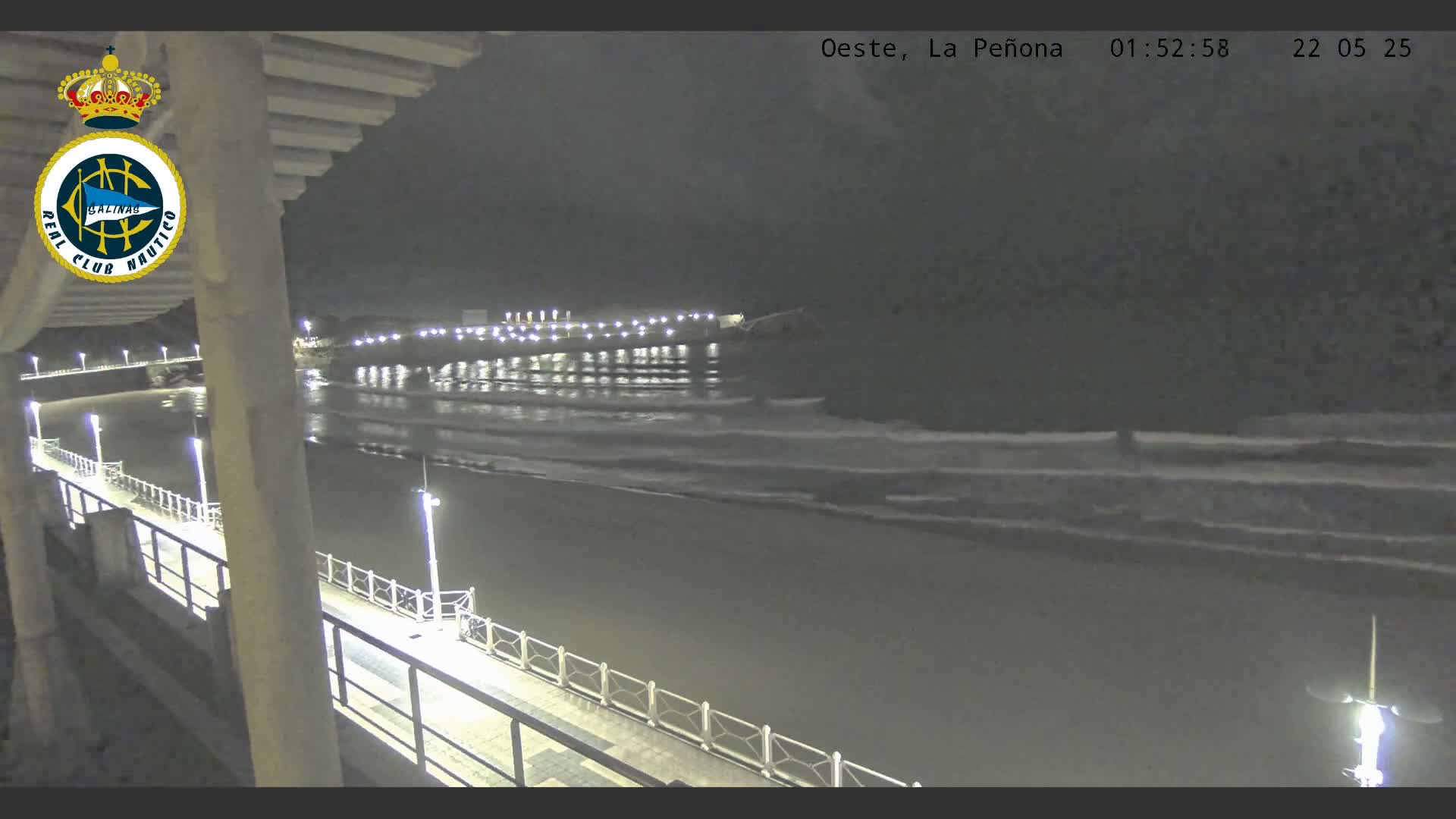 A nighttime view from an elevated position shows a pier extending over a dark beach with gentle waves lapping the shore under a dark sky.