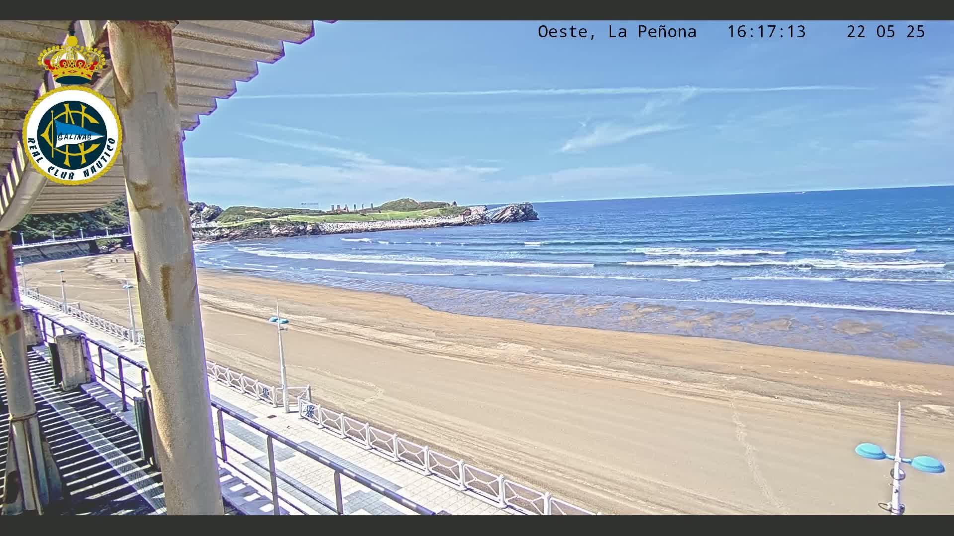A sandy beach with gentle waves under a clear blue sky is viewed from an elevated position, showing a boardwalk and a part of a building.