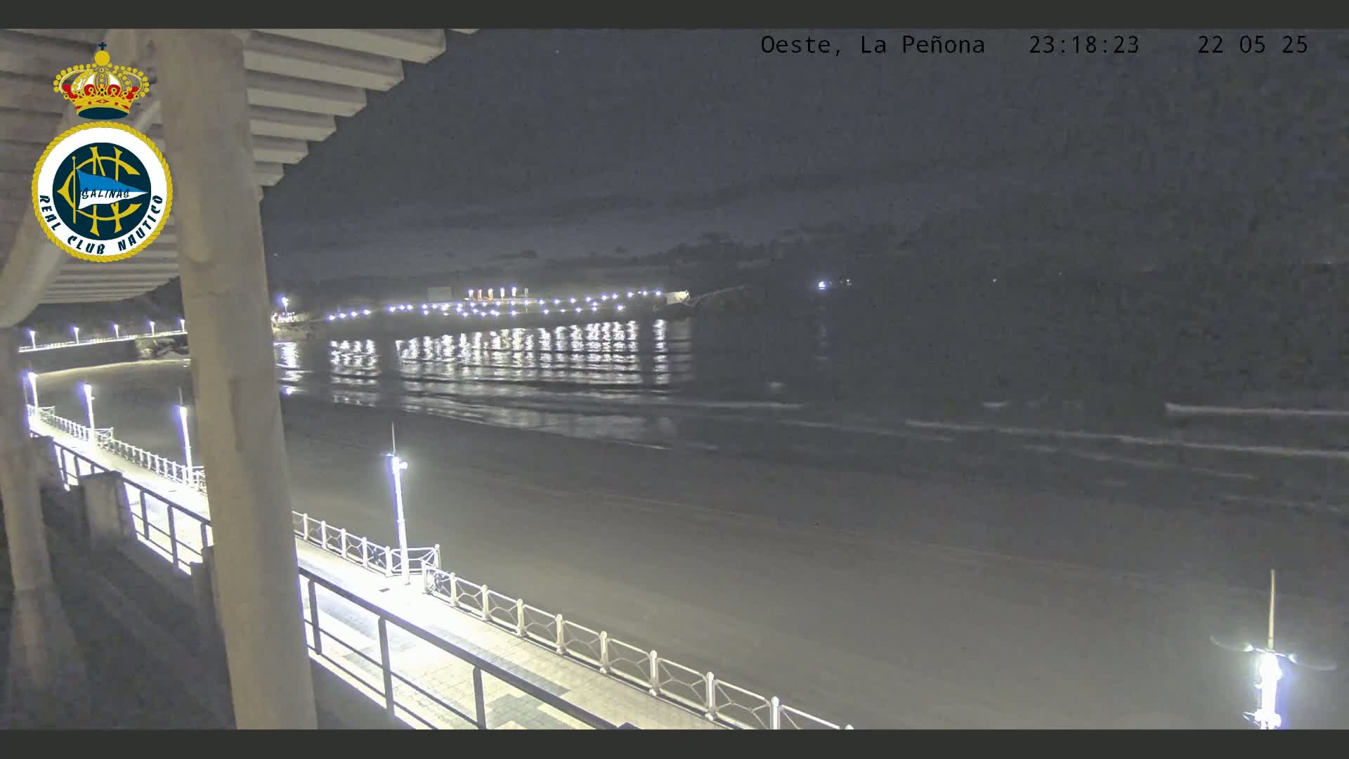 A nighttime view of a mostly empty beach with gently lapping waves, a pier extending into the water, and lights illuminating the scene.