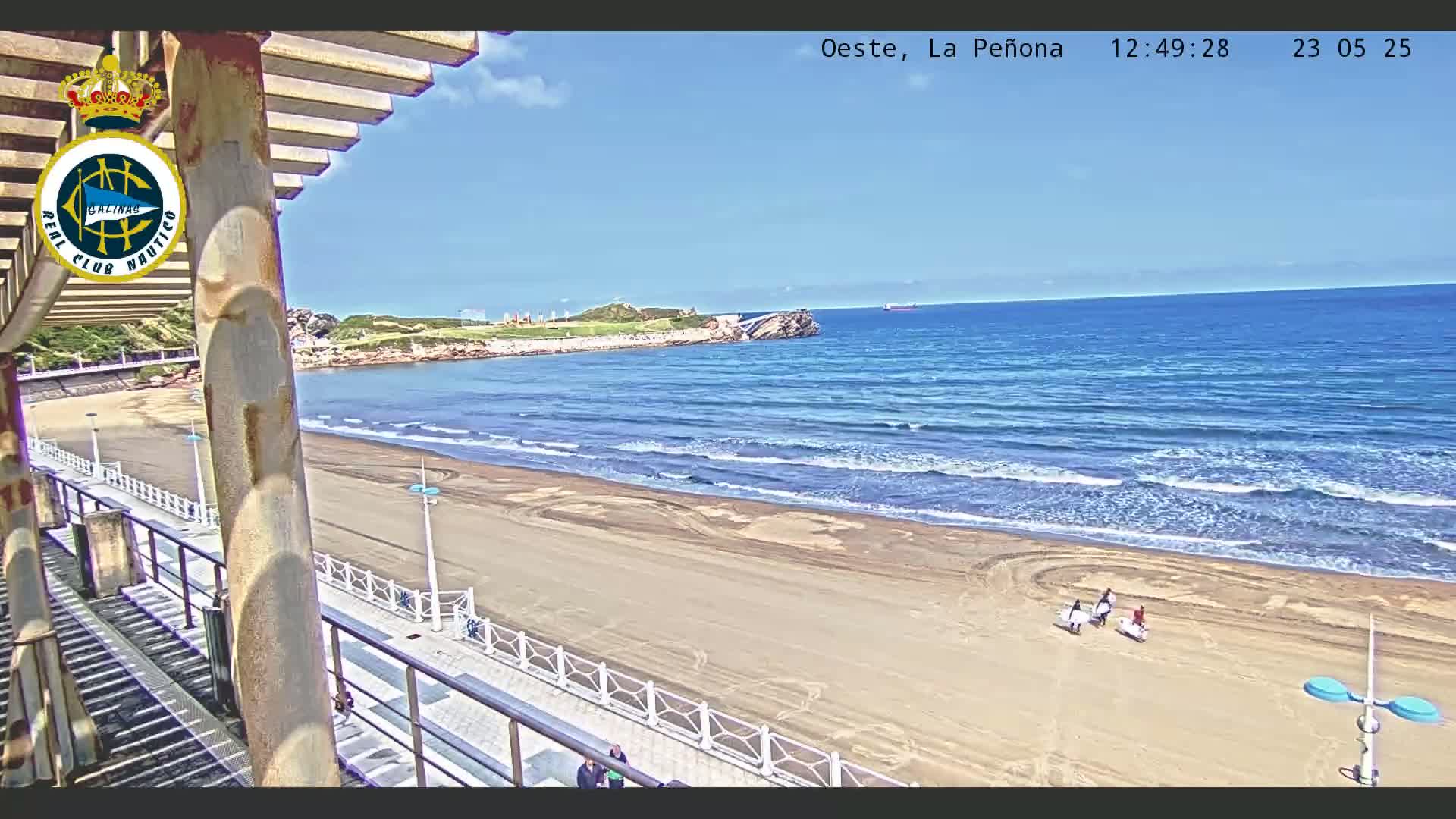 A sandy beach with a few people walking along the shore, under a mostly sunny sky, with gentle waves rolling into the ocean.